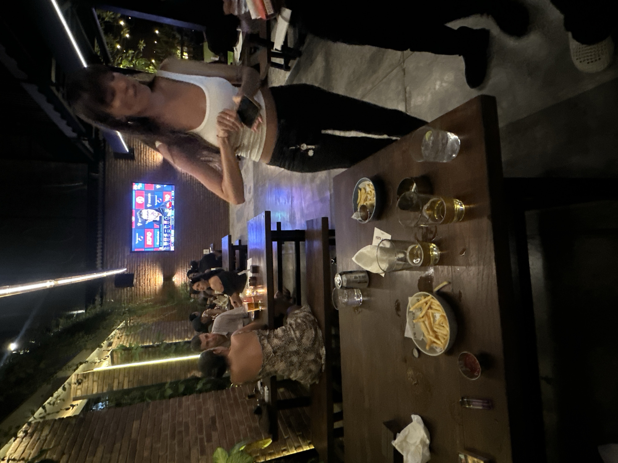 The image depicts an indoor seating area, likely a restaurant or bar, in Kuta Selatan, Indonesia. The environment features exposed brick walls, wooden tables and benches, and ambient lighting with overhead fluorescent tubes and string lights. A large television screen displaying sports programming is visible on the wall.
In the foreground, a wooden table is set with several items. A white bowl filled with french fries and a small dish of ketchup are prominently displayed. Two glasses, one containing a light-colored beverage and the other with water and a lemon slice, are also present. A can, a cigarette lighter, and crumpled napkins are scattered on the table.
In the midground, a woman with long dark hair, wearing a white tank top and black pants, is standing and holding a mobile phone. She is looking down at the phone, with her left hand extended. A keychain with keys hangs from her pants. Another person, partially visible, is standing to her right.
Further back, a couple is seated at a table. The woman is wearing a patterned dress, and the man is wearing a grey shirt. They appear to be engaged in conversation. Other patrons are visible in the background, seated at tables. The overall atmosphere suggests a casual dining or social gathering.