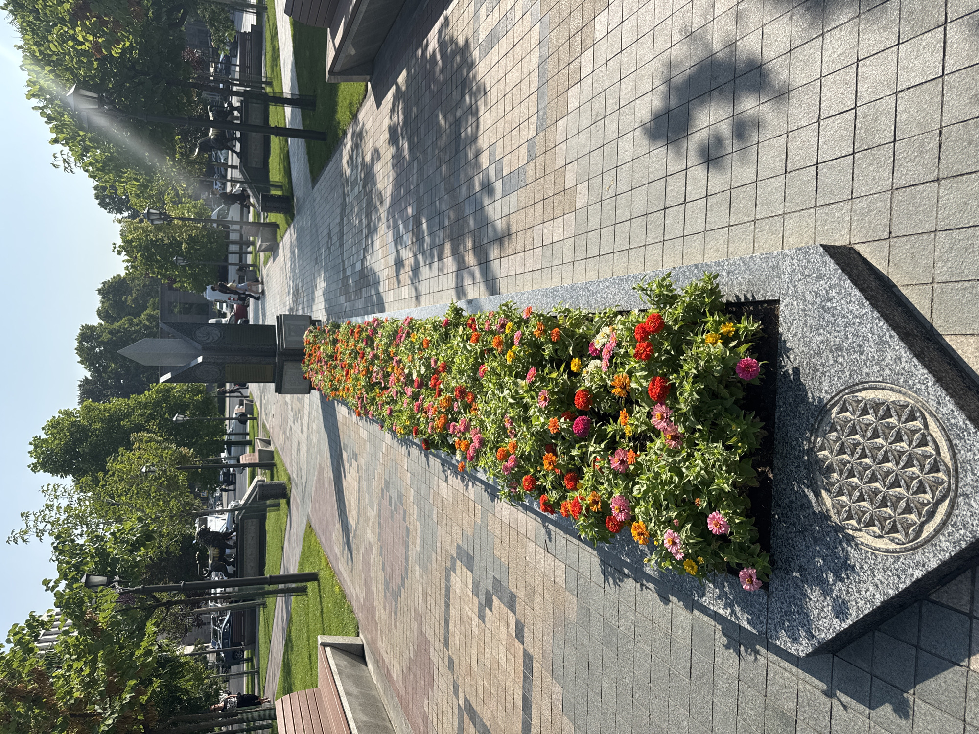 This is a photograph taken in Yerevan, Armenia, likely in a park or public square. The foreground is dominated by a long, raised flower bed filled with a variety of colorful flowers, including pink, red, and orange zinnias. The flower bed is bordered by dark granite, and on the side facing the viewer, there is a circular relief carving of the "Flower of Life" symbol embedded in the granite.
A paved walkway with a patterned design of beige, pink, and grey tiles runs alongside the flower bed. Sunlight casts dappled shadows of trees onto the walkway. In the mid-ground, more trees line the path, interspersed with lampposts and benches. Beyond the trees, there are statues of lions and bulls, and in the distance, cars and people are visible, suggesting a public area within the city. A large, dark monument with intricate carvings stands at the end of the flower bed, near the walkway's intersection. The sky is clear and blue, indicating a sunny day.