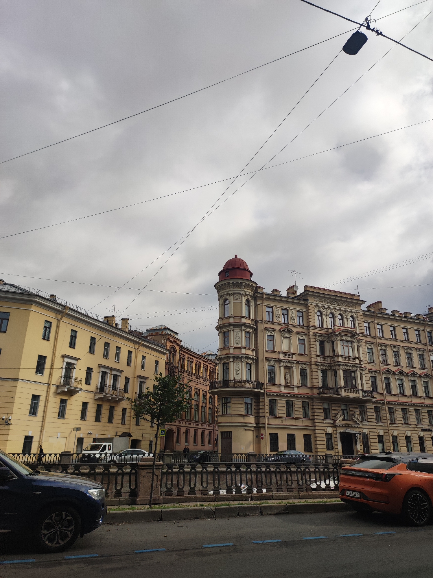 This is a medium-angle, slightly tilted, horizontal exterior, daytime, overcast, street-level shot of buildings and cars in Saint Petersburg, Russia. The view is looking down a canal or river with a decorative metal railing along the bank. Two multi-story buildings dominate the frame. The building on the right is a cream-colored, ornate structure with many windows and balconies, topped with a red domed turret on the left side of its facade. The building on the left is a lighter yellow with fewer ornate details, also featuring multiple stories and balconies. A light-colored van and several other cars are parked or driving along the street in front of the buildings. A dark blue SUV is parked in the foreground on the left, and an orange coupe is parked on the right, facing away from the camera. Overhead, electrical wires crisscross the cloudy, gray sky, with a single light fixture suspended from one of the wires. The general impression is of an urban environment in an older European city.