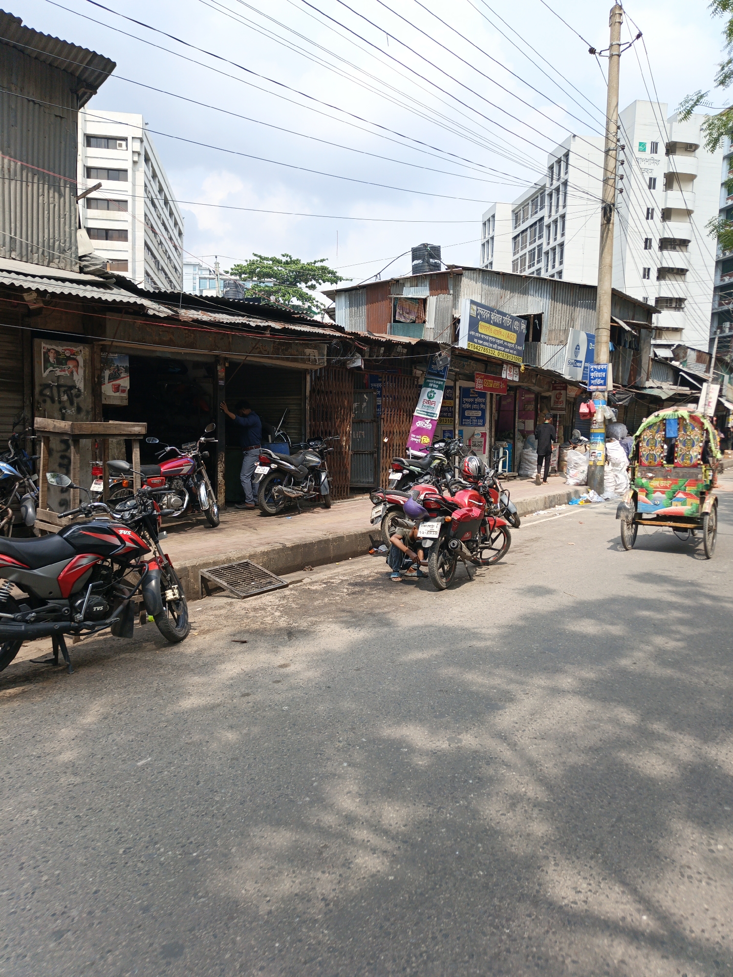 The image captures a street scene in an urban area, possibly in South Asia, with a blend of commercial and residential elements. The photograph is taken from street level, showing the asphalt road surface in the foreground, with shadows cast by trees overhead.
Along the side of the road are several parked motorcycles, some with their kickstands down. Adjacent to the road is a line of shops or businesses, characterized by corrugated metal roofing and rudimentary storefronts. Some businesses are adorned with signs and banners, with a prominent sign for "Sundarban Courier Service" in Bengali script. People are visible engaging in activities around the shops, with one person appearing to be working on a motorcycle.
In the background, taller buildings rise, including a white structure with multiple floors. The buildings suggest a densely populated urban environment with a mixture of old and new infrastructure. Power lines are strung across the sky.
A colorful rickshaw is also present on the right side of the image, adding to the diversity of transportation options. The environment has a workaday atmosphere.