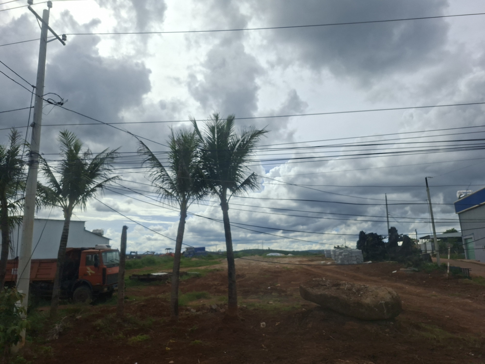 The image depicts a construction site on a cloudy day in Thôn 9, Vietnam. In the foreground, a large, oblong rock rests on reddish-brown dirt. To the left, an orange dump truck is parked near a white building. Several palm trees are scattered throughout the scene, some partially obscuring the view. In the middle ground, stacks of white materials are visible, along with dark, possibly tarpaulin-covered, objects. The background shows a hilly landscape with more construction activity, including structures and what appears to be a crane. Overhead, numerous power lines crisscross the sky, which is dominated by dark, heavy clouds. The overall impression is one of an active, possibly developing, area.