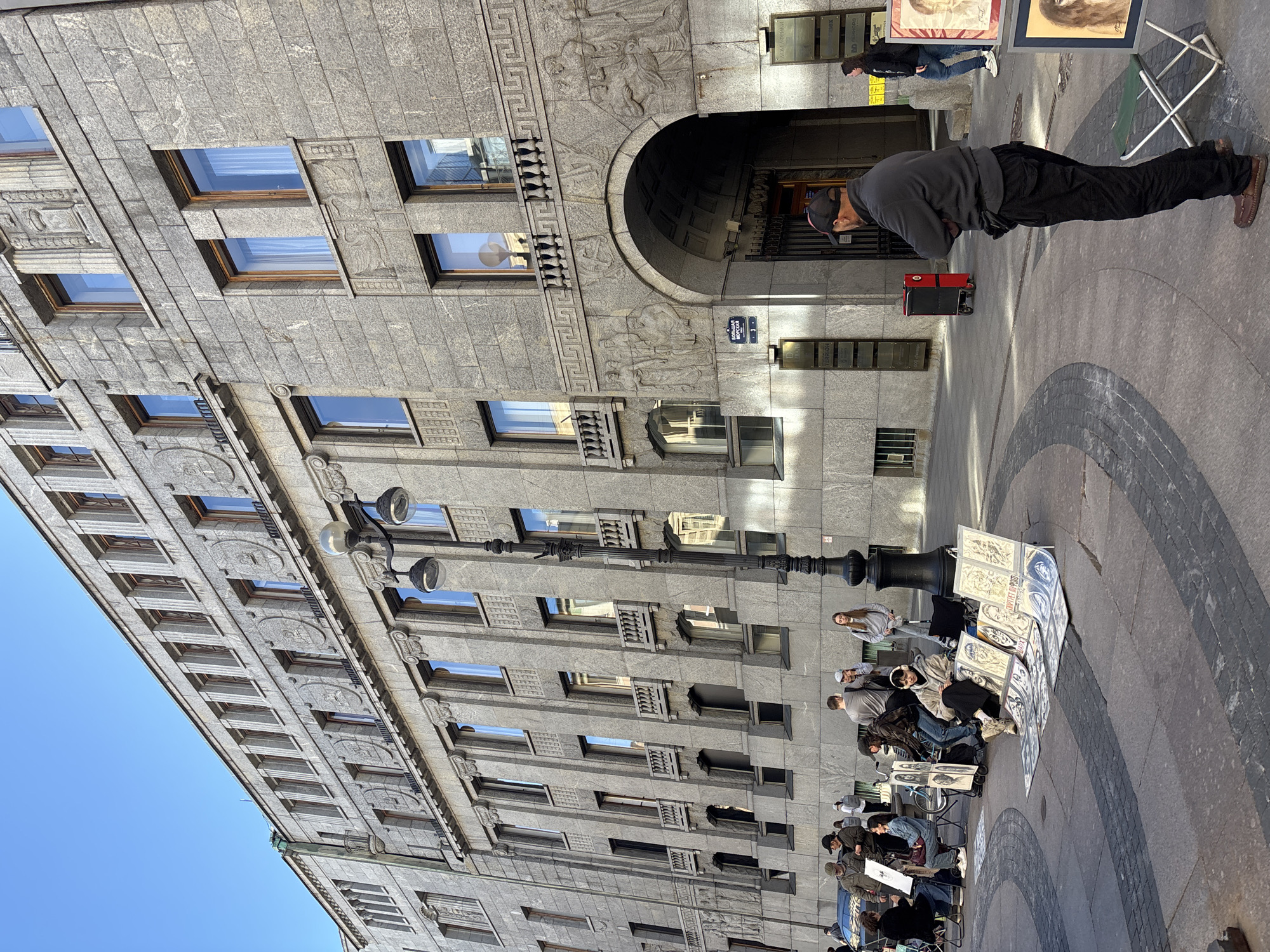 The image shows a street scene in Saint Petersburg, Russia. The focus is on a large, ornate stone building with numerous windows. In the foreground, several people are gathered, some seated and others standing. A man in a dark grey hoodie and black pants is standing near the building's entrance, looking down at something in his hands. To his right, a collection of framed artworks, seemingly portraits, are displayed on an easel. A folding chair is set up next to the display.
Further down the street, other individuals are interacting, and there appears to be an artist or vendor interacting with potential customers around the artwork. The ground is paved with stone, featuring a circular pattern. A lamppost is visible on the side of the building, and street signs are mounted on the wall. The sun is casting shadows, suggesting it is daytime, likely late morning or afternoon. The sky is clear and blue. The overall atmosphere appears to be a bustling urban environment with an artistic element. Visible text includes "БОЛЬШАЯ МОРСКАЯ 3" on a street sign.