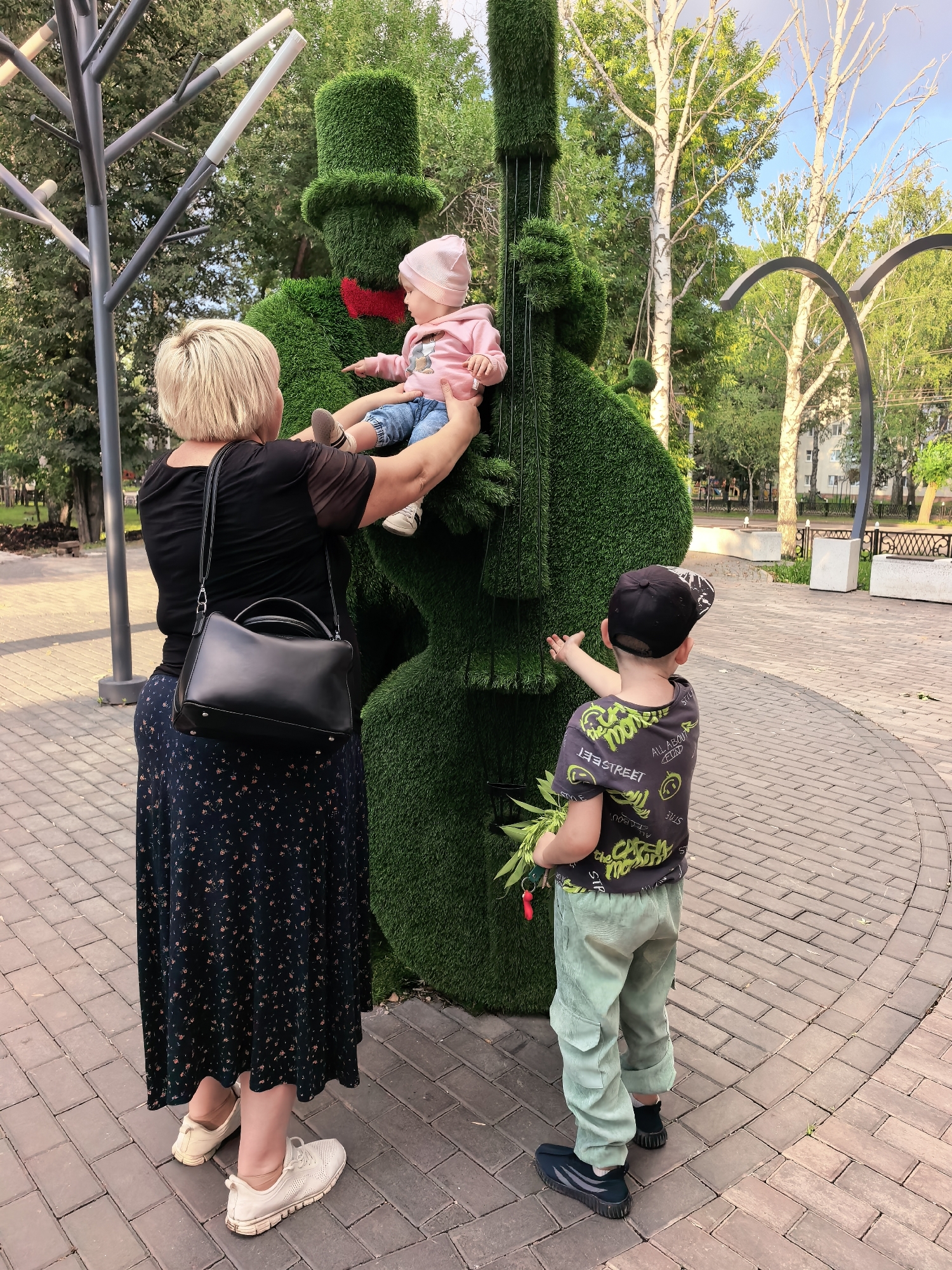 In Sterlitamak, Russia, a woman with blonde hair is holding a baby wearing a pink outfit and a pink hat, pointing towards a large, green, grass-covered sculpture shaped like a cello with a red bow tie and a top hat. To the right of the sculpture, a young boy wearing a black baseball cap, a grey t-shirt with green lettering, and light green pants is reaching out to touch the sculpture. He is holding a small bunch of green leaves. The scene takes place on a brick-paved area with trees and park benches in the background, under a clear blue sky.
