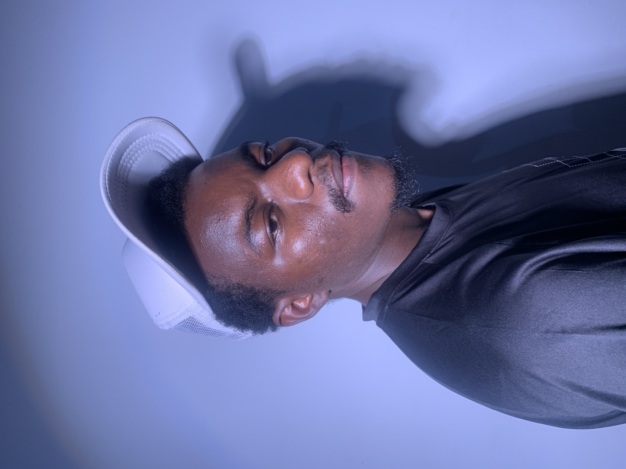 A close-up, medium shot portrait of a young Black man, from the chest up, is captured against a plain, light blue background. The man is wearing a white mesh trucker hat tilted slightly to the left and a black t-shirt. He is looking off to his right, with his head turned slightly away from the camera. The lighting creates a dramatic shadow of his head and shoulder on the wall behind him. His skin is slightly oily, and he has a mustache and a goatee. The background is an unadorned wall. The scene is likely an indoor studio or a room with controlled lighting. There is no indication of a specific city or location.