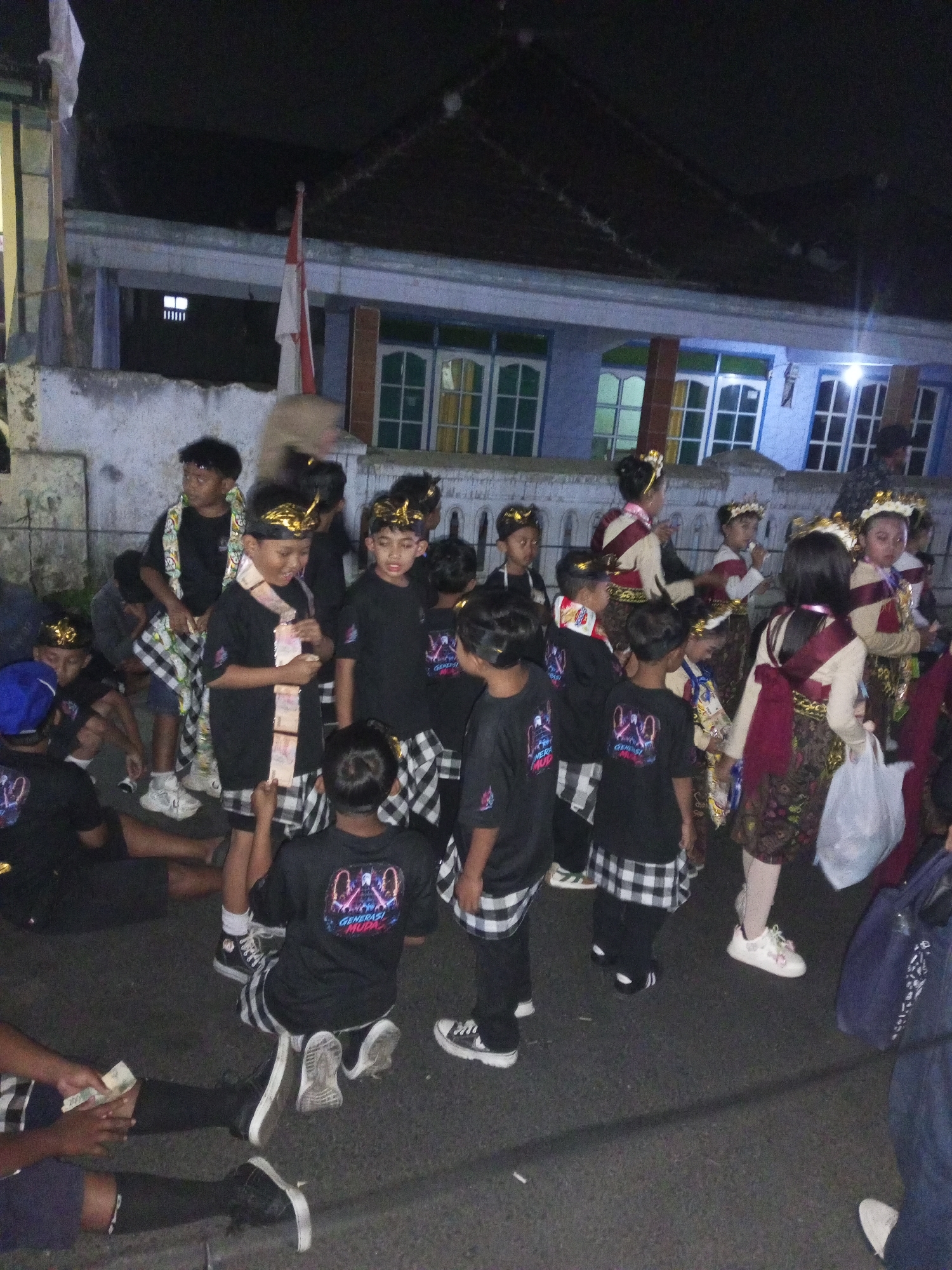 In Langlang, Indonesia, a group of children, dressed in traditional Balinese attire, are gathered outdoors at night. The children, some wearing black t-shirts with colorful graphics and checkered skirts, others in elaborate headdresses and sashes, stand and sit on a paved surface in front of a building with a blue facade. Some of the children are holding or interacting with what appear to be money or small bundles. A red and white flag is visible in the background, suggesting a local or national event. The lighting suggests artificial illumination, possibly from streetlights or nearby buildings, creating a somewhat dim and shadowed atmosphere. The overall scene appears to be a performance or a gathering related to a cultural event.