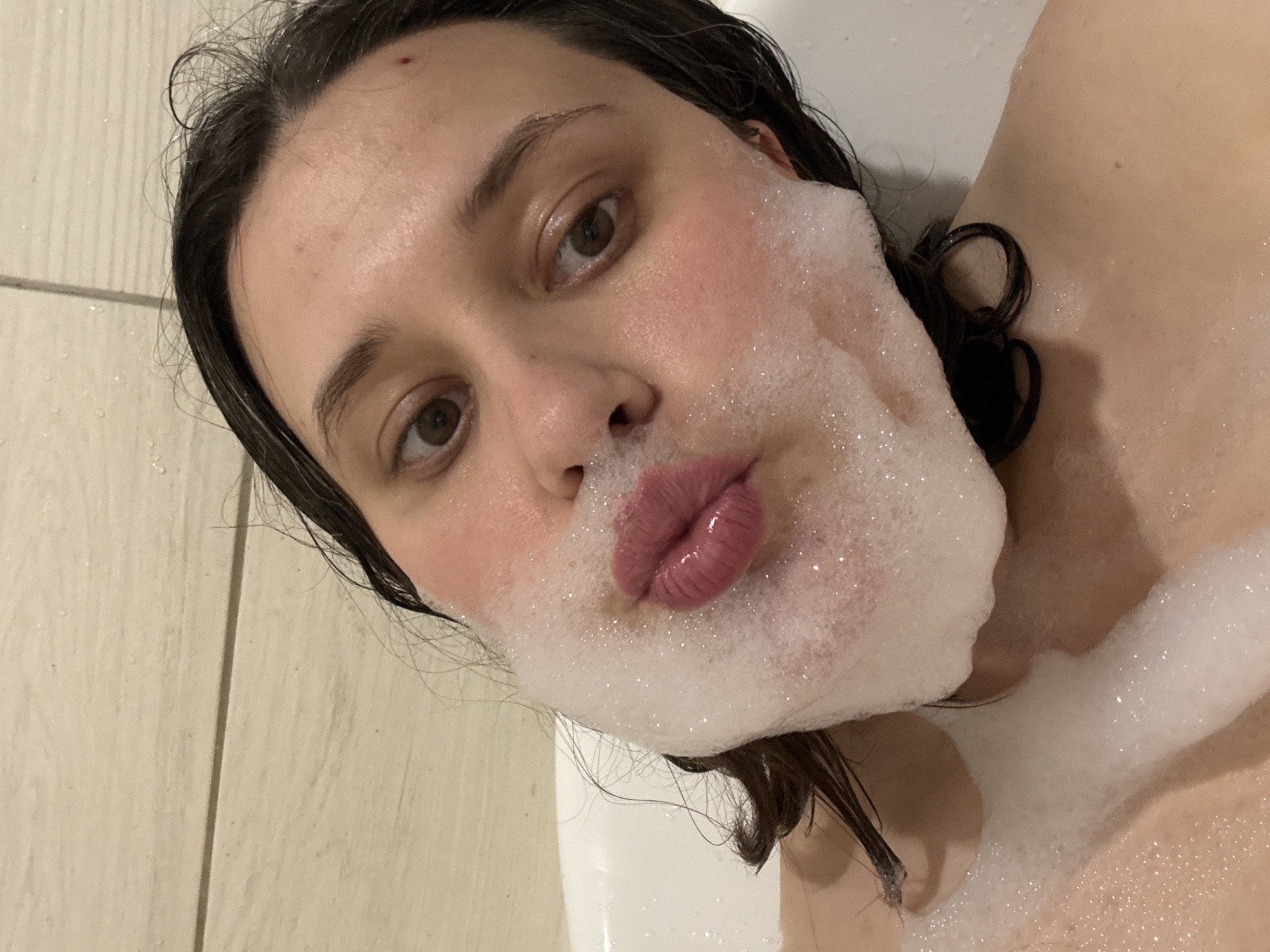 A person with dark hair and fair skin is lying in a bathtub, covered in soap bubbles. The person's face is visible, with a slight pout. The background shows a beige tile wall and the white surface of the bathtub. The location is unknown.