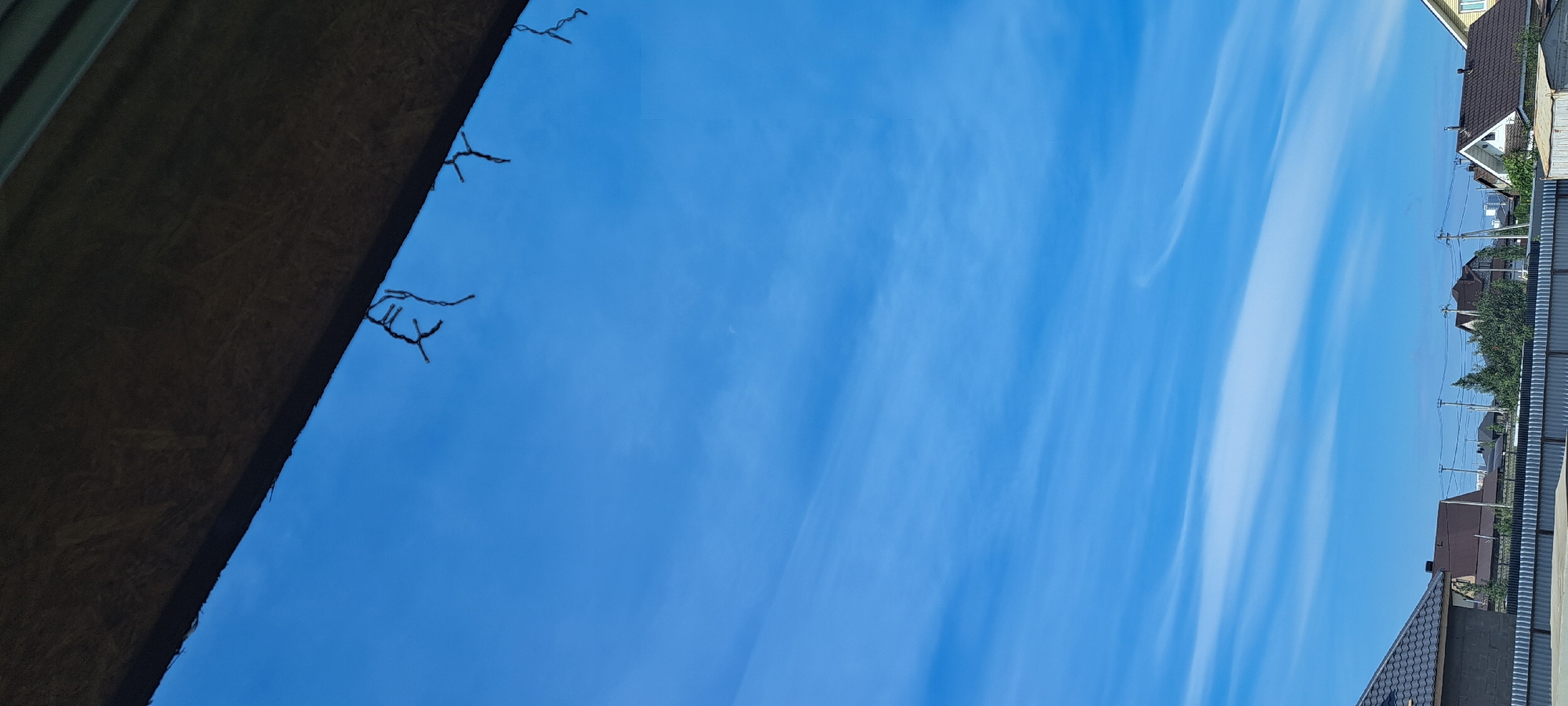 The image shows a clear blue sky with thin, wispy clouds stretching horizontally across the frame. A dark, textured overhang, possibly a roof or awning, occupies the top portion of the image, with thin, dark strands hanging down from it. Below the overhang, the sky is the dominant feature, with a faint crescent moon visible in the upper left quadrant. In the lower portion of the image, a residential area is visible. Several houses with pitched roofs, some with dark shingles, are seen behind a metallic fence. Green trees are interspersed among the houses, and utility poles with wires are visible. The scene appears to be a residential neighborhood in the daytime, possibly the village of Az'mushkino, Russia, given the presence of houses and the rural feel. There are no people or specific actions depicted in the image.