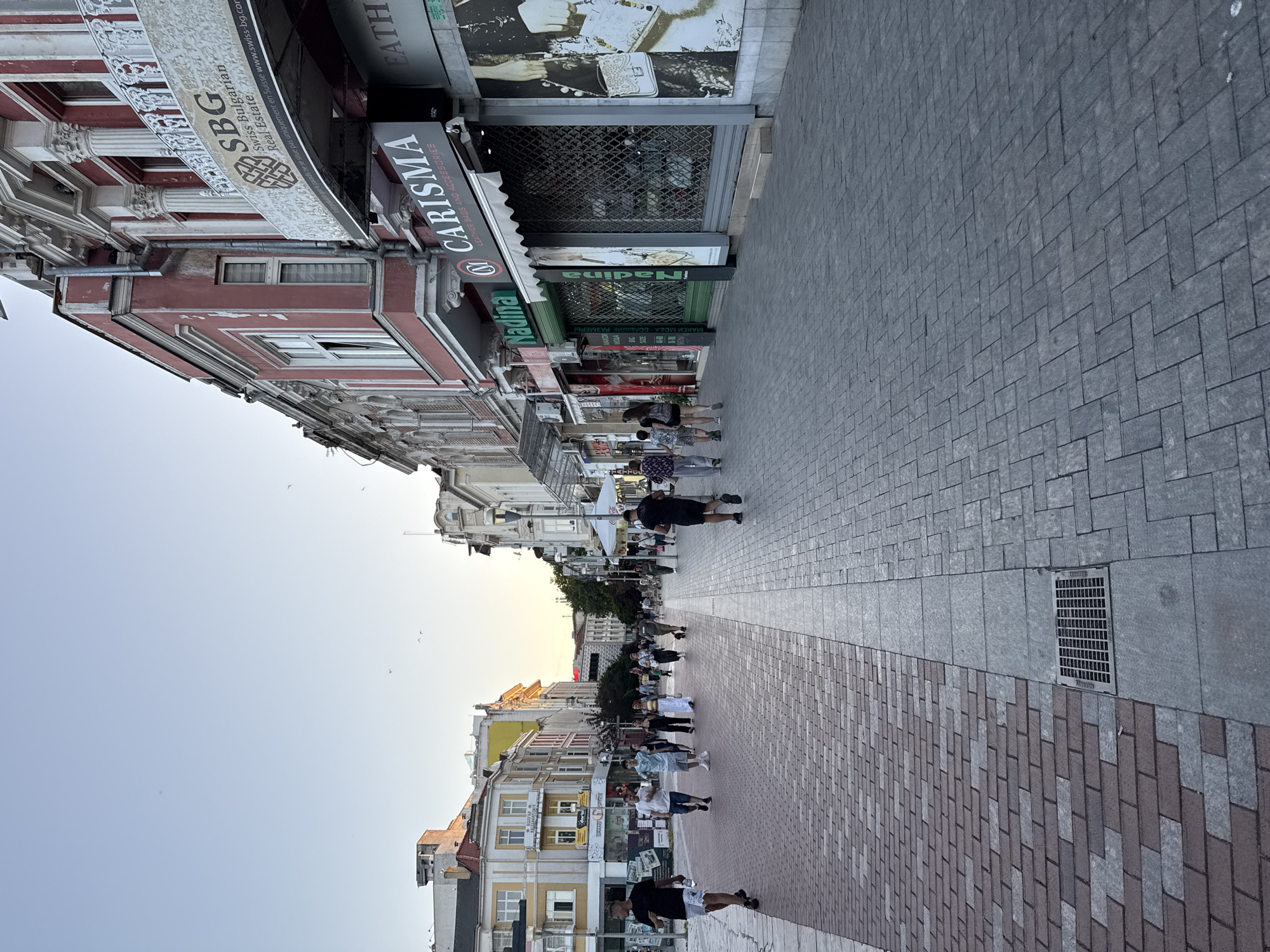 A street scene depicts a wide pedestrian walkway paved with alternating bands of reddish-brown and gray interlocking bricks. A metal drain grate is visible on the walkway. Buildings line the right side of the street, displaying classical architecture with decorative facades, balconies, and cornices. Several shops are identified by signage, including "SBG Swiss Bulgarian Real Estate," "Nadina," and "CARISMA LEATHER BAGS AND ACCESSORIES." The buildings on the left side of the street are also multi-story structures with varied architectural styles and window designs. A clear, pale blue sky with a few distant birds suggests it is either early morning or late afternoon. Several people are walking along the pedestrian area in various directions, some individually and some in small groups. The overall atmosphere is that of a commercial street in a European city.