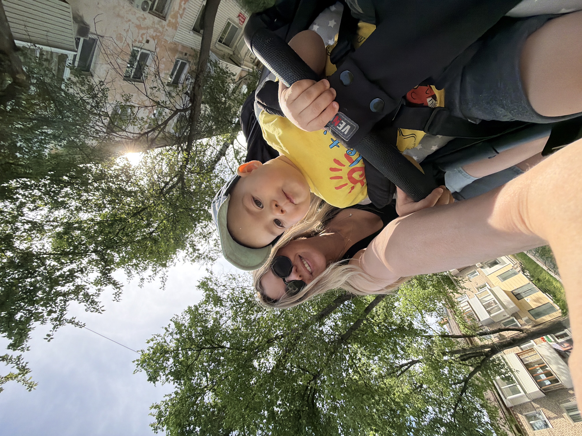 A low-angle outdoor shot shows a blonde woman in sunglasses holding the handle of a stroller, in which sits a young child in a yellow shirt and a blue cap. The woman smiles at the camera. The child looks up at the camera. In the background, foliage and buildings are visible. The image appears to be taken in Chaikovsky, Russia.