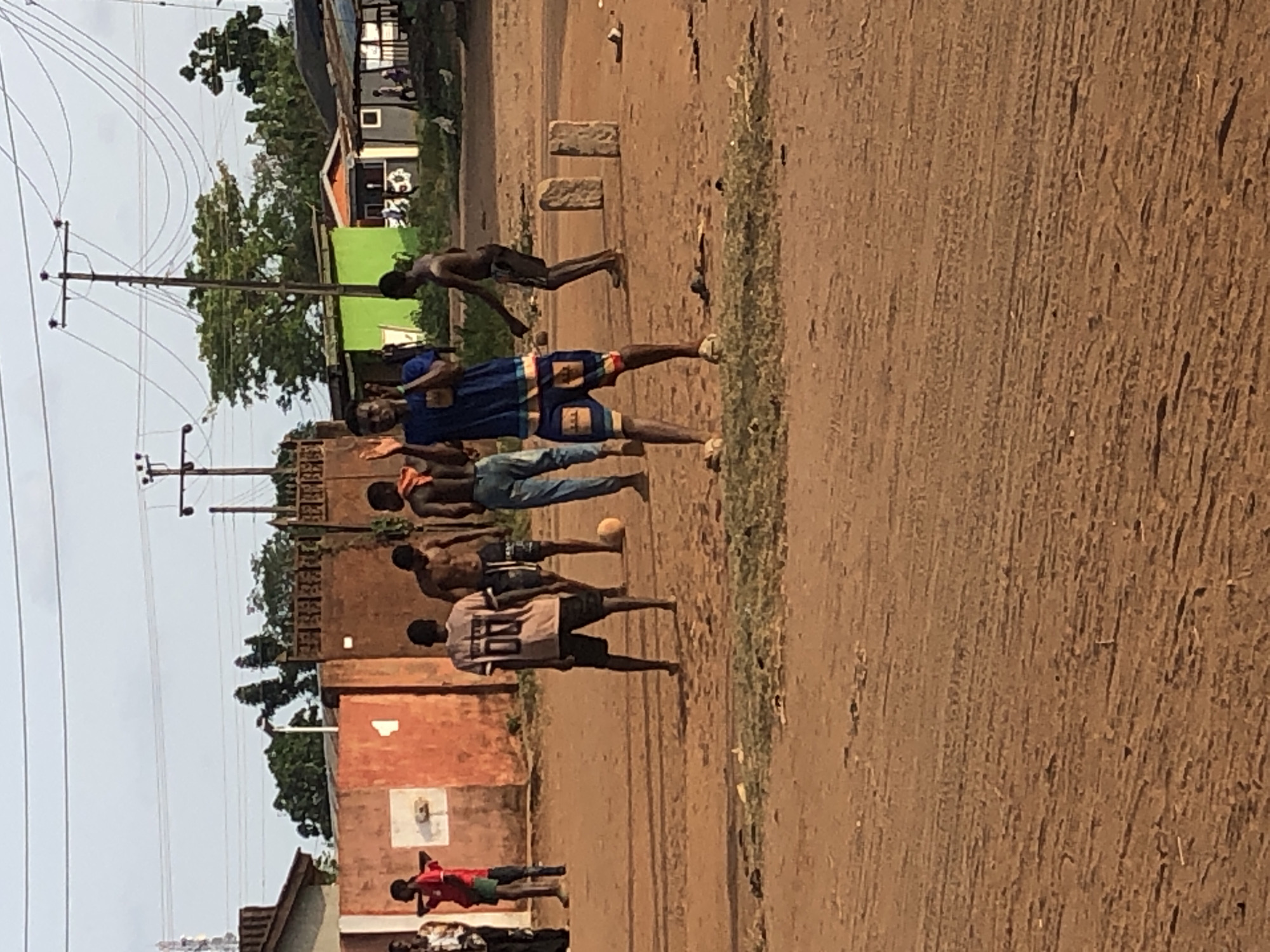 A street scene in Unknown, Unknown shows a group of about seven boys and men engaged in a street soccer game, with a ball visible near the center of the group. The scene includes a building with a basketball hoop on the wall, utility poles, a dirt road, and sparse greenery.