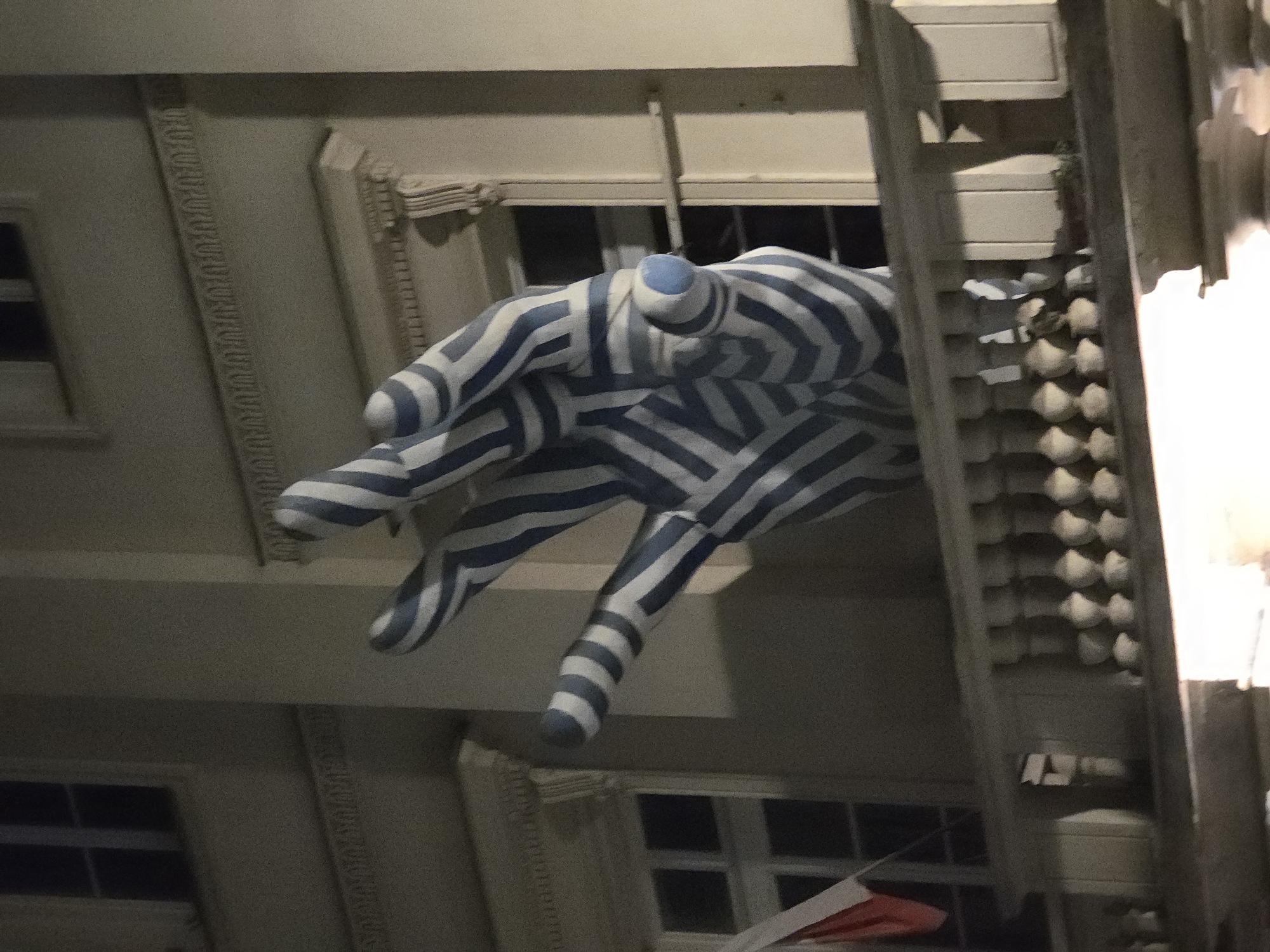 Captured from a low angle in Warsaw, Poland, the scene features a large, sculpture of a hand painted with blue and white stripes. The hand appears to be suspended from a building, with architectural details such as windows, molding, and a decorative cornice visible in the background. The surrounding buildings suggest an urban environment. The scene is illuminated, likely at night.
