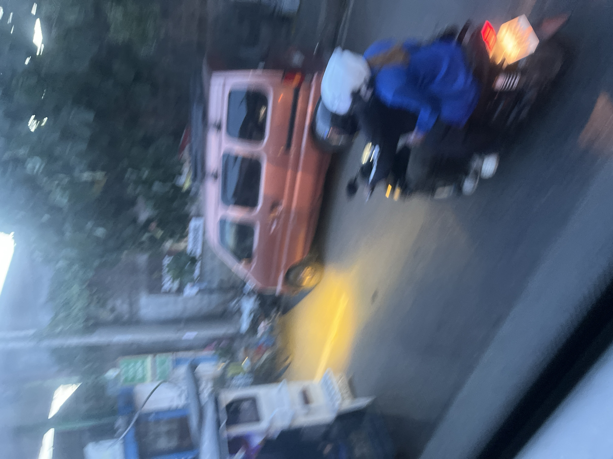 The image shows a street scene in Davao City, Philippines, possibly during the evening or at dusk, as indicated by the artificial lighting and the muted colors of the surroundings. The primary focus is on a small, light pink van parked on the side of a road. In front of the van, two people are on a motorcycle. The person in front is wearing a white helmet, and the person behind is wearing a blue jacket. They appear to be stationary, possibly interacting with the van or waiting.
The van has tinted windows and a roof rack, suggesting it might be used for transportation or carrying goods. The street appears to be paved, and there are hints of buildings and greenery in the background, though the overall image is blurry, making precise identification difficult. A strong yellow light, likely from a street lamp or the van's headlights, illuminates a portion of the road.
There are other objects visible on the side of the road, including what appear to be some sort of wheeled carts or structures, possibly for vendors or storage. Some green signage is also present, but the text is illegible due to the blurriness. The overall impression is one of a typical urban street in the Philippines, with everyday activities taking place. The blurriness suggests the photo was taken while in motion or with a low-quality camera, or has been subjected to image processing that has reduced its sharpness.