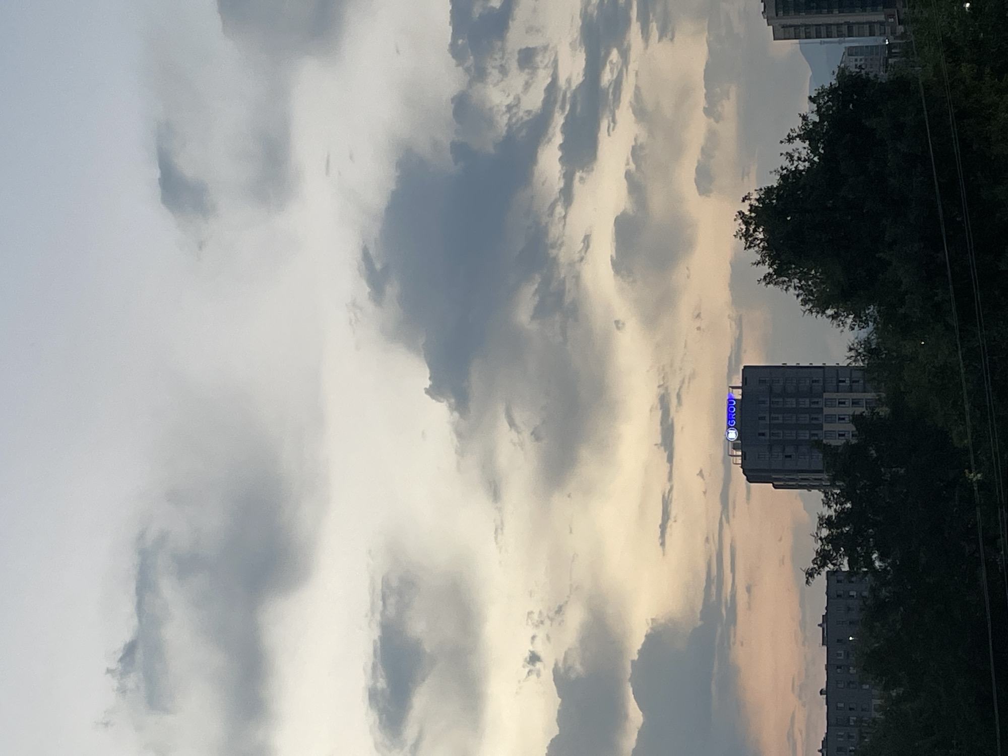 The image shows a slightly overcast sky with fluffy clouds in muted shades of gray and white, transitioning to a warm, pale yellow near the horizon, suggesting either dawn or dusk. In the middle ground, a tall, modern building stands prominently, its facade a mixture of light-colored panels and darker windows. At the very top of this building, a blue neon sign with white lettering reads "GROU" within a circular logo. To the left of this building, another, lower, more uniform building with many windows is visible. In the foreground, a dense line of dark green trees stretches horizontally across the lower part of the image, partially obscuring the lower sections of the buildings. In the far right background, the silhouette of a mountain range is faintly discernible against the sky. This scene appears to be an urban landscape in Almaty, Kazakhstan, likely taken from a residential area overlooking the city. There are no people or animals visible in the image.