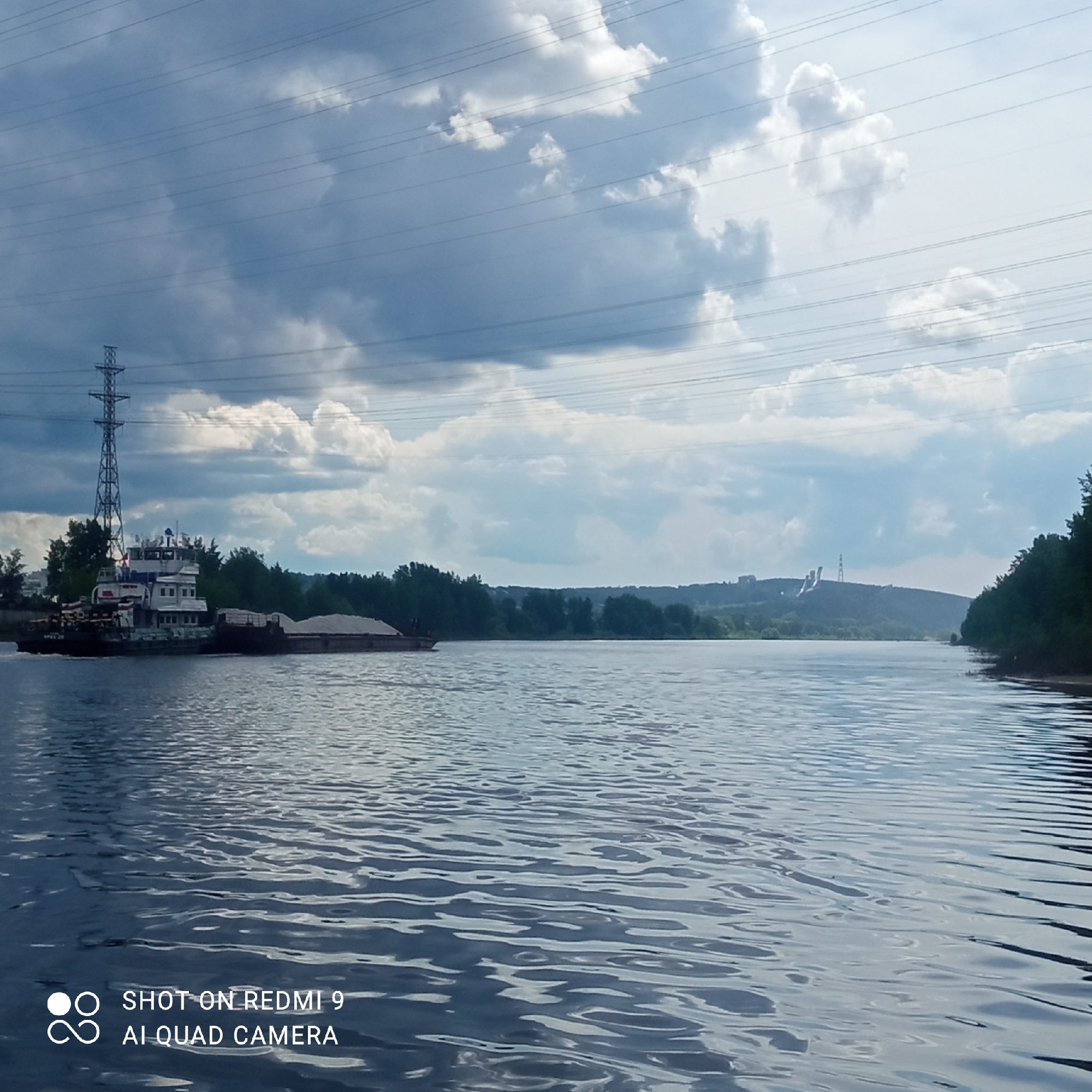 The image depicts a river scene, presumably in the city of Chaikovsky, Russia, given the provided information. A tugboat is pulling a barge loaded with material, possibly gravel or sand, on the river. An electrical transmission tower is visible on the left bank, near the tugboat. Across the river, the landscape includes a line of trees along the bank, a hill in the distance, and structures atop the hill, including what appears to be a ski jump. The sky is partially cloudy. The water surface shows gentle ripples. The text "SHOT ON REDMI 9 AI QUAD CAMERA" is in the lower left corner, indicating the image was captured with a Redmi 9 phone.