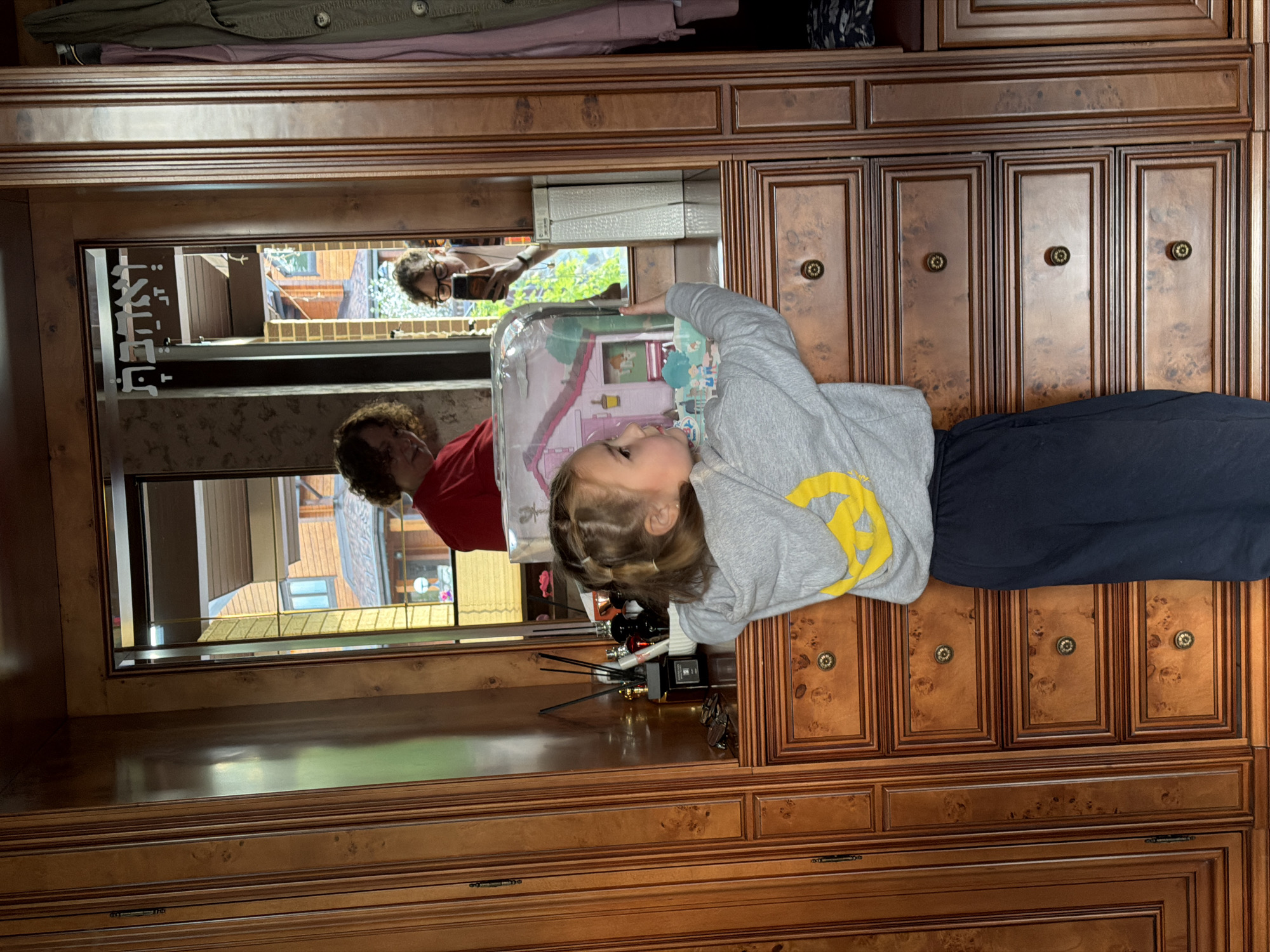A little girl in a gray shirt and dark pants lies on a wooden dresser with a dollhouse in a clear plastic container. The dresser has a built-in mirror that reflects an adult female in a red shirt and a person holding a phone, along with a view of buildings outside. The scene is in Новоглаголево, Russia.