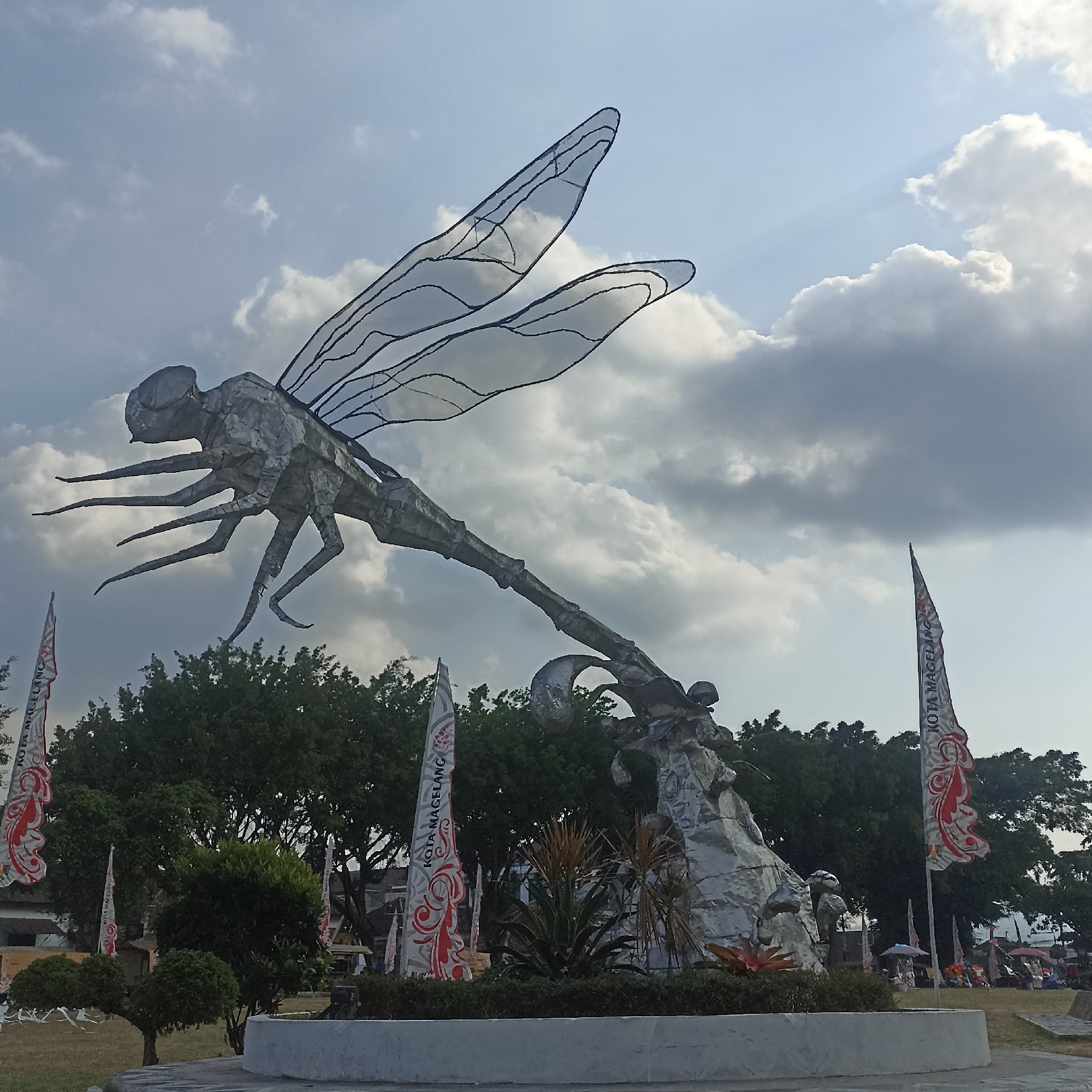 The image depicts a large metal sculpture of a dragonfly, with its wings outstretched, against a cloudy sky. The dragonfly is positioned prominently in the foreground. In the background, there are trees, several flags with decorative patterns and text that reads "KOTA MAGELANG", and a scattering of people and market stalls. The sculpture appears to be located in an outdoor public space.