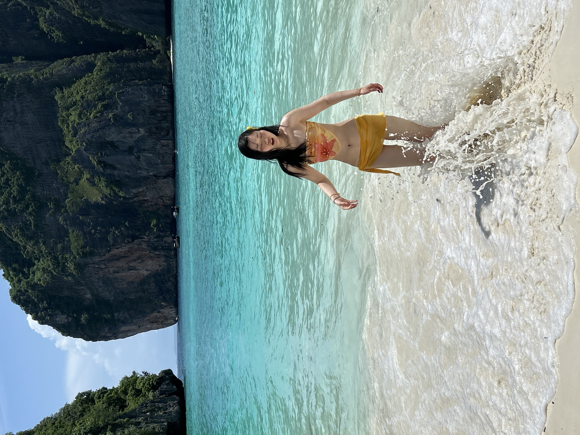A young Asian woman stands in shallow, turquoise water on a sandy beach. She is wearing a yellow bikini top with a floral pattern and a matching yellow sarong tied around her waist. Her eyes are closed, and she is looking down as she walks forward, creating a splash of water around her legs. The background features dramatic limestone cliffs covered in lush green vegetation, characteristic of the Ao Nang region in Krabi Province, Thailand. The water is clear and calm, with small waves lapping at the shore. The sky is blue with scattered white clouds.