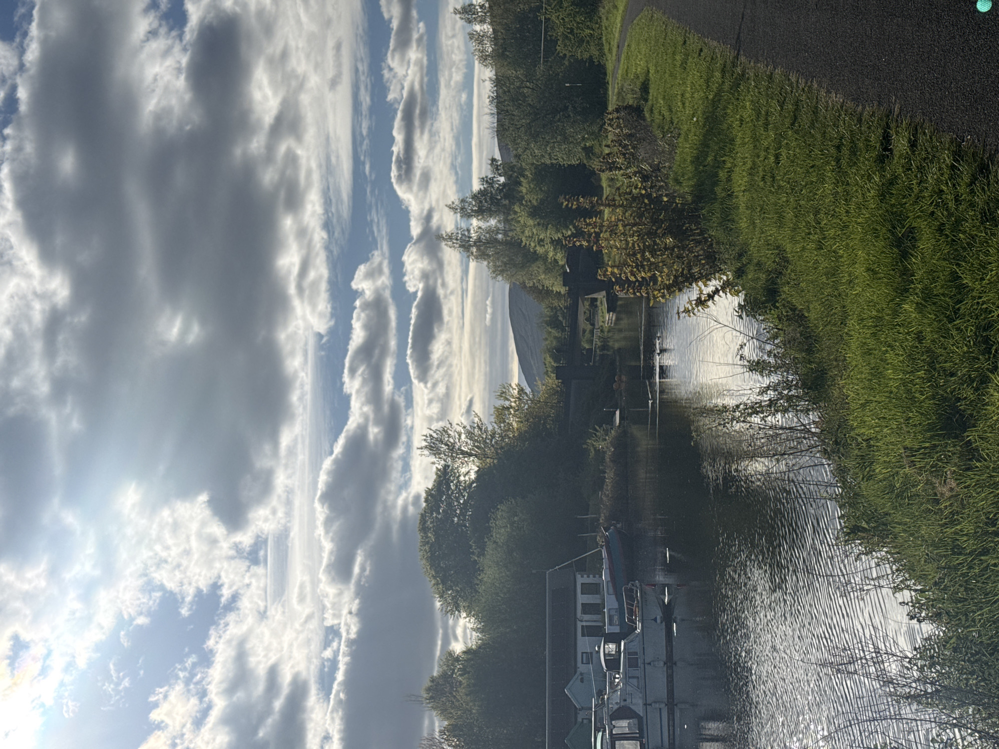 The image depicts a waterway scene in an unknown city and country. The sky above is filled with mostly gray clouds with the sun shining through in sections. A canal runs parallel to a grassy bank and a paved path. There are trees growing along the bank, next to boats. Ripples in the water and the reflections indicate slight movement.