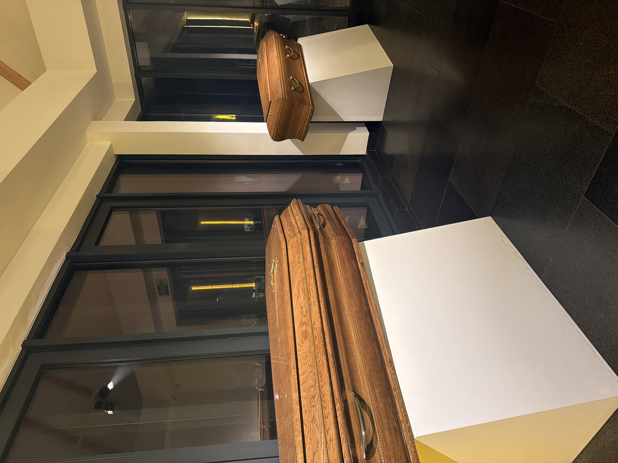 The image shows an indoor exhibition space, likely a gallery or museum. The primary subjects are two wooden coffins displayed on white pedestals. The larger coffin in the foreground is richly detailed with a cross and ornate metal handles, while the smaller one in the background is simpler in design. The background features a wall of dark-framed windows reflecting interior lights. The floor is covered in dark, patterned tiles. No people are visible. The location is given as Unknown, Unknown.