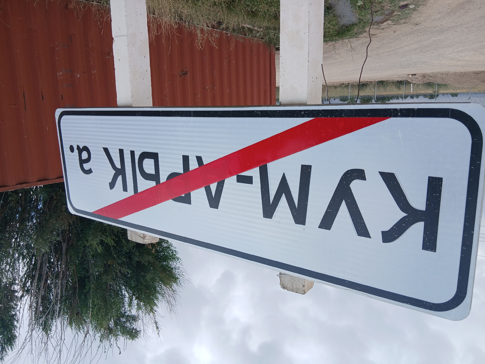 A road sign indicating the location of Kum-Aryk, Kyrgyzstan is pictured. The sign is white with a black border and black lettering. A red diagonal stripe is overlaid on the sign, indicating that the location is no longer within the city limits. The sign is supported by two concrete posts. The background shows a cloudy sky, trees, a fence, and a dirt road. A corrugated metal wall is visible behind the sign.