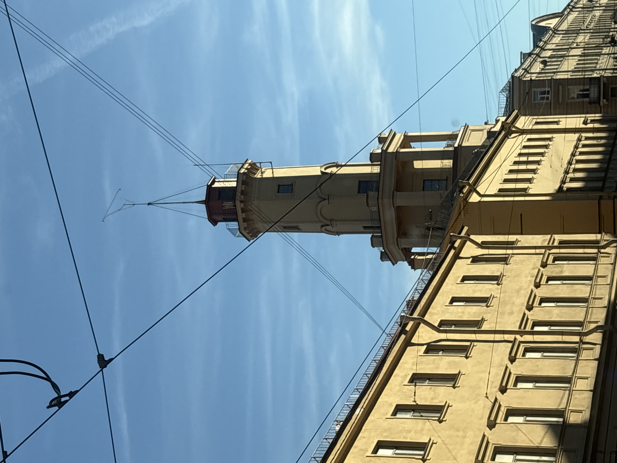 The image shows a tall, slender tower with a red top, possibly a lighthouse or observation tower, rising from a pale yellow building with many windows. The building appears to be in an urban setting, with other buildings visible in the background and overhead wires crisscrossing the sky. The sky is a clear, bright blue with thin white clouds. The lighting suggests it is daytime, likely late morning or early afternoon, with the sun casting shadows on the buildings. The scene is shot from a low angle, looking upwards towards the tower and the sky. The overall impression is of a grand, old European city. There is no visible text in the image. The image is oriented vertically, with the tower appearing to stand upright in the center.