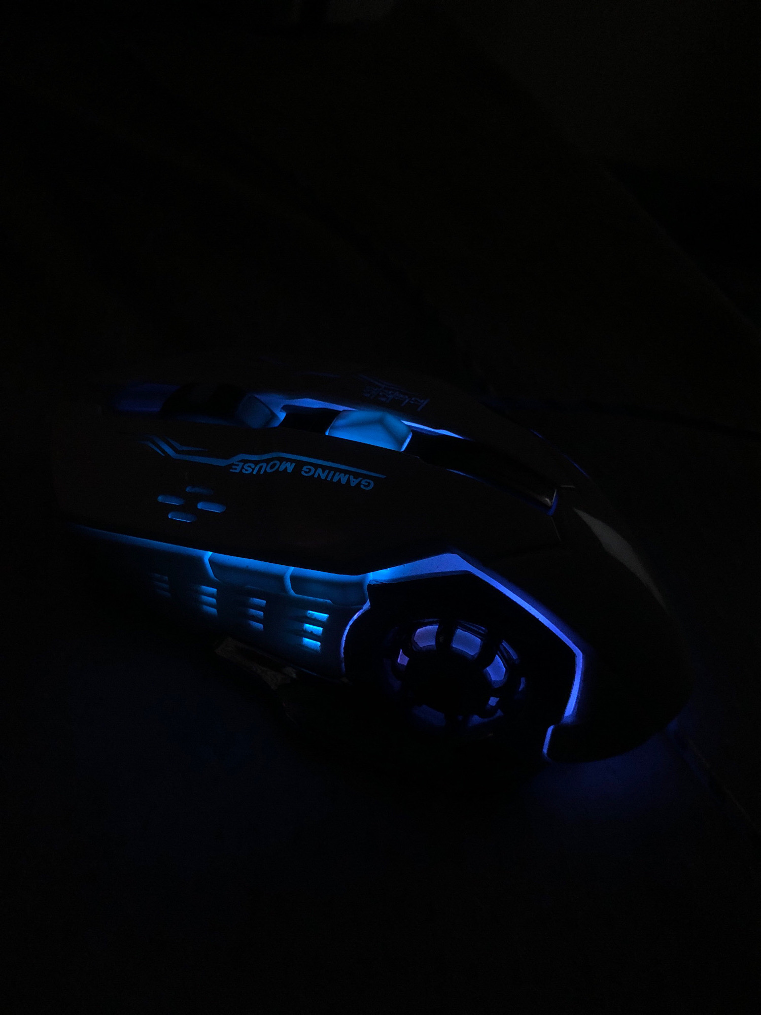 A dark, low-angle close-up photo shows a black gaming mouse with illuminated blue accents. The mouse is positioned on a dark surface, likely a desk or mousepad, with the front-left portion of the mouse most visible and illuminated. The word "GAMING MOUSE" is printed on the side of the mouse in light blue text, oriented vertically and reversed. The scroll wheel area and the back of the mouse also emit a soft blue light, highlighting its angular, aggressive design. The overall environment is dark, with the only light sources being the mouse's LEDs. There are no people or other discernible objects in the frame, and no information can be derived about Saidan, Pakistan from this image.