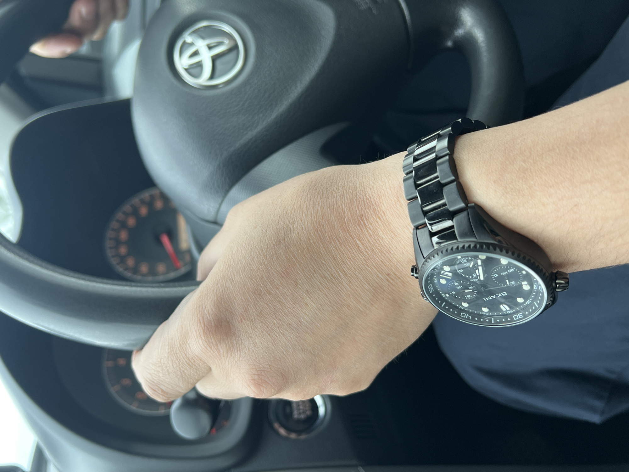 A first-person perspective shot shows a driver's left hand, adorned with a black wristwatch, gripping the steering wheel of a car. The dashboard, featuring a Toyota emblem, is visible in the background. The scene is indoors, possibly within a vehicle, and no specific location in Yuzhno-Sakhalinsk, Russia, can be identified from the image.