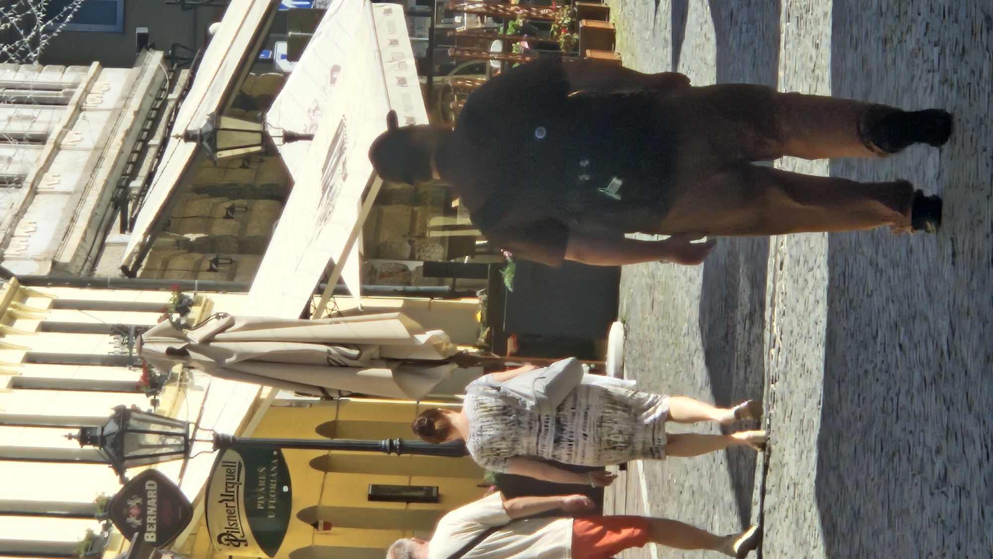 The image depicts a street scene in Prešov, Slovakia, with a focus on individuals walking away from the camera. In the foreground, a man wearing a dark shirt and light brown trousers walks on a cobblestone street, his back to the viewer. He appears to have a backpack on. To his left, a woman in a patterned dress walks ahead, also with her back to the camera, carrying a light gray bag over her shoulder. To the far left, a man wearing a light-colored shirt and orange shorts walks in the same direction. The background features buildings with yellow and stone facades, a classic street lamp, and awnings extending from the buildings. Several potted plants and outdoor seating elements are visible, suggesting a commercial area or outdoor dining establishment. The overall lighting indicates a sunny day with distinct shadows cast on the cobblestones.