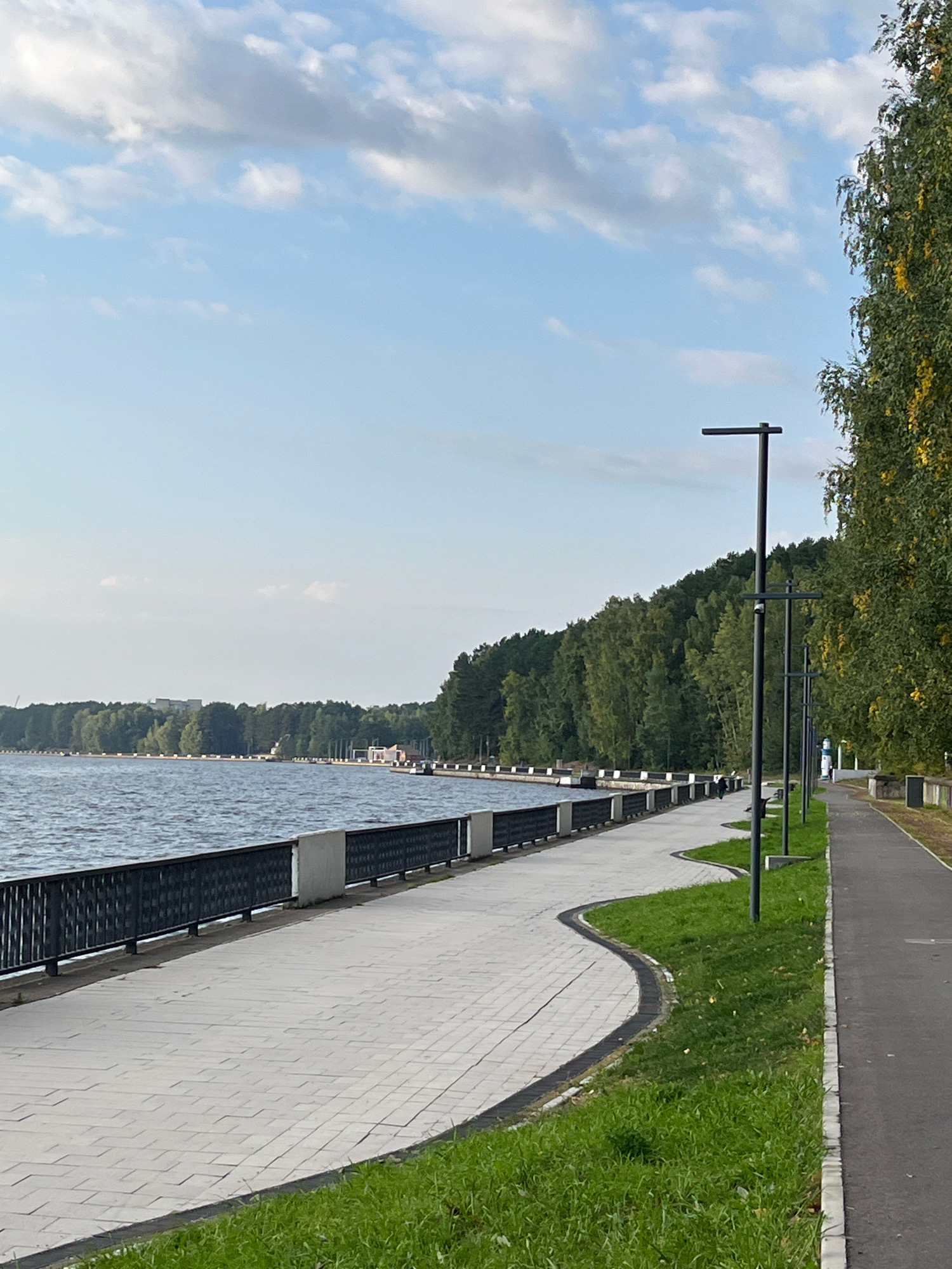The image depicts a scenic waterfront promenade in Chaikovsky, Russia. A wide, paved walkway with a patterned surface curves along the edge of a calm body of water. A dark metal railing with ornate detailing separates the walkway from the water. Beyond the water, a dense forest lines the opposite shore, with some buildings visible in the distance. On the right side of the walkway, a paved asphalt path runs parallel, bordered by a strip of green grass. Several modern black lampposts stand along the walkway, spaced at intervals. A few indistinct figures are visible walking on the promenade. The sky is a pale blue with scattered white clouds. A tall tree with yellowing leaves is visible on the far right, suggesting it might be autumn.