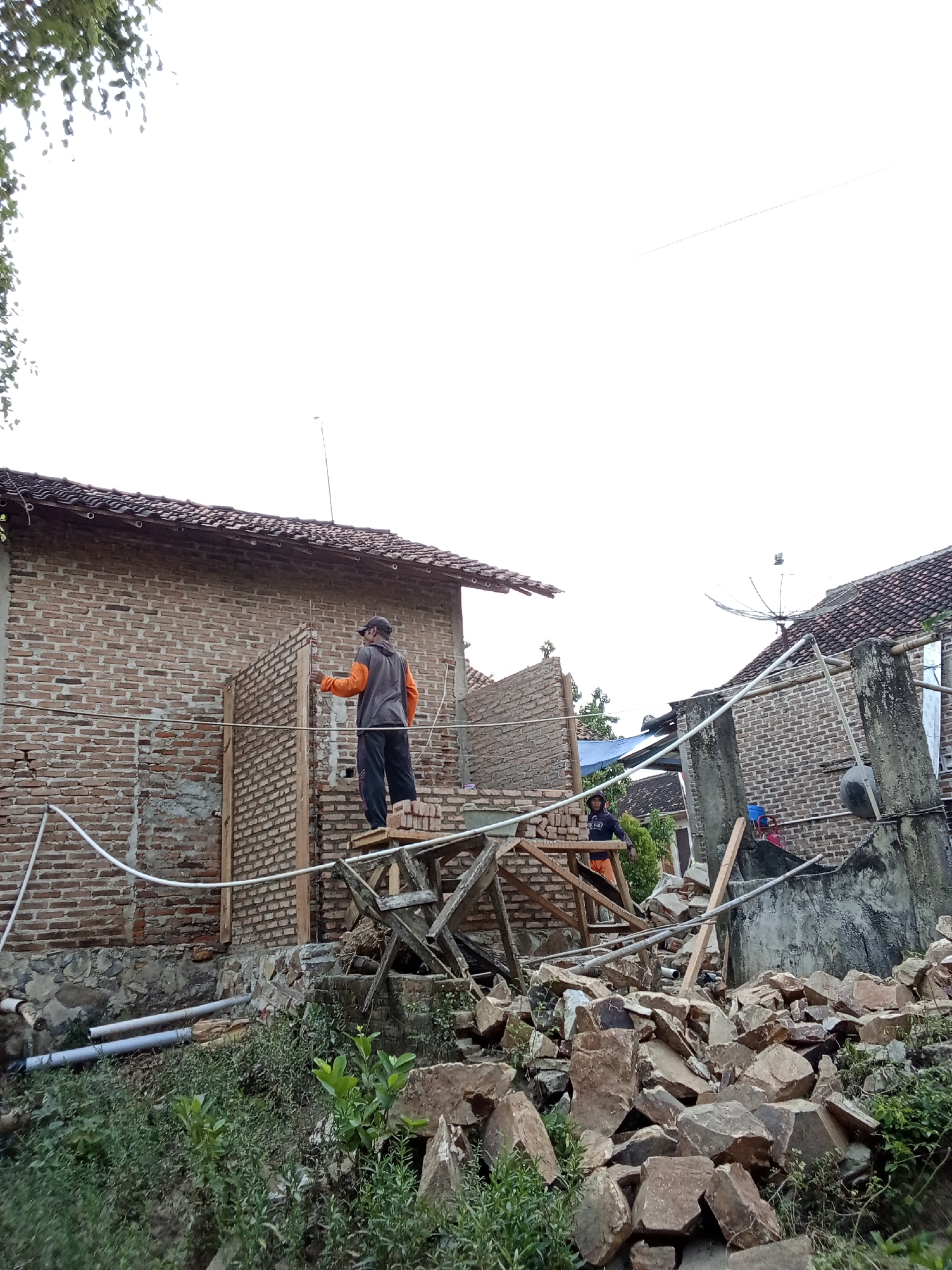 In Pringsewu Barat, Indonesia, an outdoor scene depicts a partial demolition or construction of a brick building. Two individuals are present. One person, wearing an orange jacket and dark pants, stands on a wooden scaffolding, facing and interacting with the brick wall. Another individual, in a dark shirt and orange pants, is also on scaffolding further back. The environment is characterized by scattered rubble, including large stones, debris from the building structure, and exposed brickwork. White pipes are visible running along the ground and on the building's exterior. The sky is overcast.