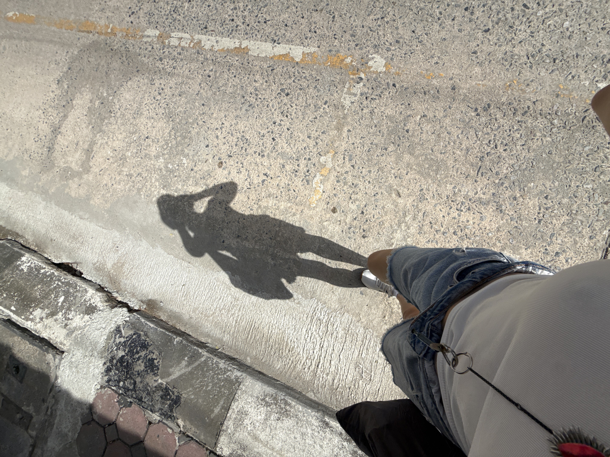 A first-person perspective shot from Karon, Thailand, shows a person's shadow cast on a concrete street. The shadow depicts the individual holding a camera, suggesting they are taking a selfie. The person is wearing ripped denim shorts and a white tank top. The street has a yellow and white painted line marking. To the left, there is a concrete curb and a sidewalk made of interlocking hexagonal pavers. The lighting indicates it is daytime.