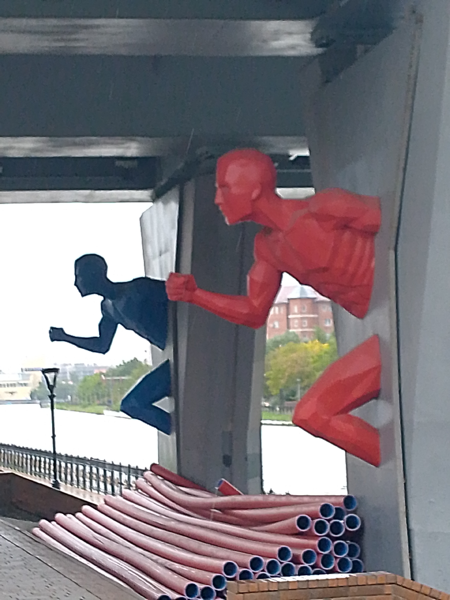 The image shows two abstract sculptures of runners, one in red and one in dark blue, emerging from a grey concrete structure, possibly a bridge or an underpass. The red runner is in the foreground and on the right, with its torso and legs visible. The dark blue runner is behind and to the left, also depicted in a running pose. In the background, a body of water, a railing, a lamppost, and trees are visible, suggesting an outdoor location, possibly a waterfront promenade. A stack of red, flexible pipes is in the lower right portion of the image. The overall scene appears to be in an urban environment. The prompt specifies the location as Astana, Kazakhstan.