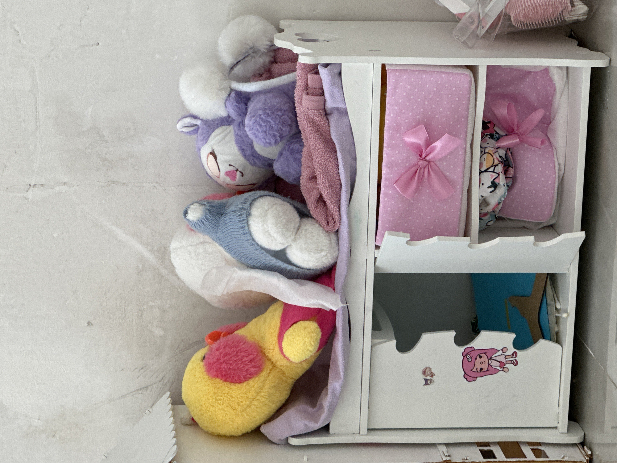 A white wooden dollhouse/storage unit is situated against a white wall. The top shelf contains pink folded items with bows, and a pink hairbrush in a clear plastic wrapper. The middle shelves contains towels and stuffed animals, including a purple toy with white pom-poms, a blue-and-white item, and a yellow-and-pink plush toy. The bottom shelf holds books and is decorated with cartoon character stickers. The location is an indoor setting of Unknown, Unknown.