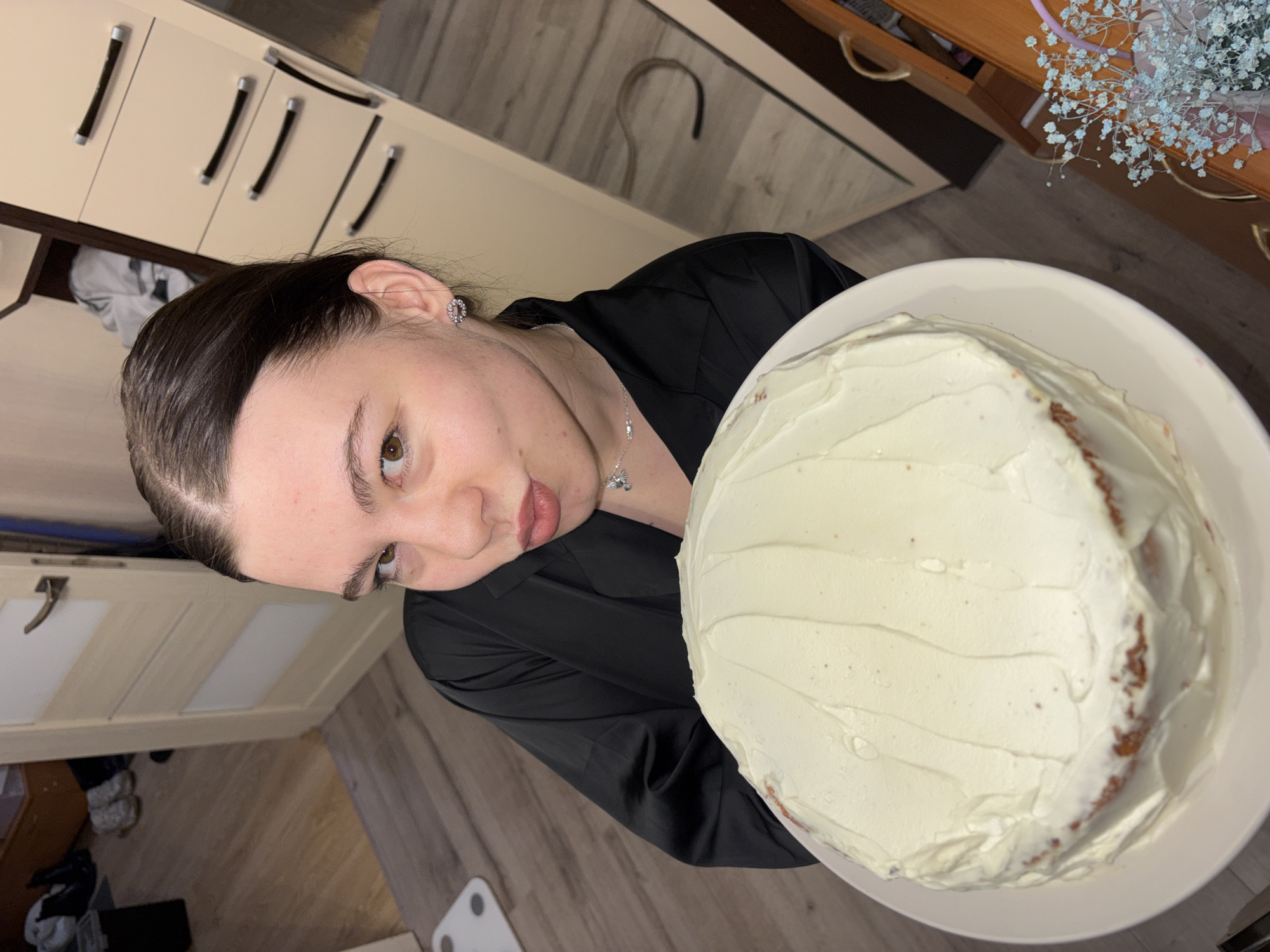 A person is holding a round cake with white frosting in a white plate. The person has brown hair pulled back, brown eyes, and is wearing a black shirt and a silver necklace and earrings. The background includes kitchen cabinets, wooden flooring, a white scale, and a bouquet of white flowers on a wooden surface. There is no indication in the picture the location is in Perm, Russia.
