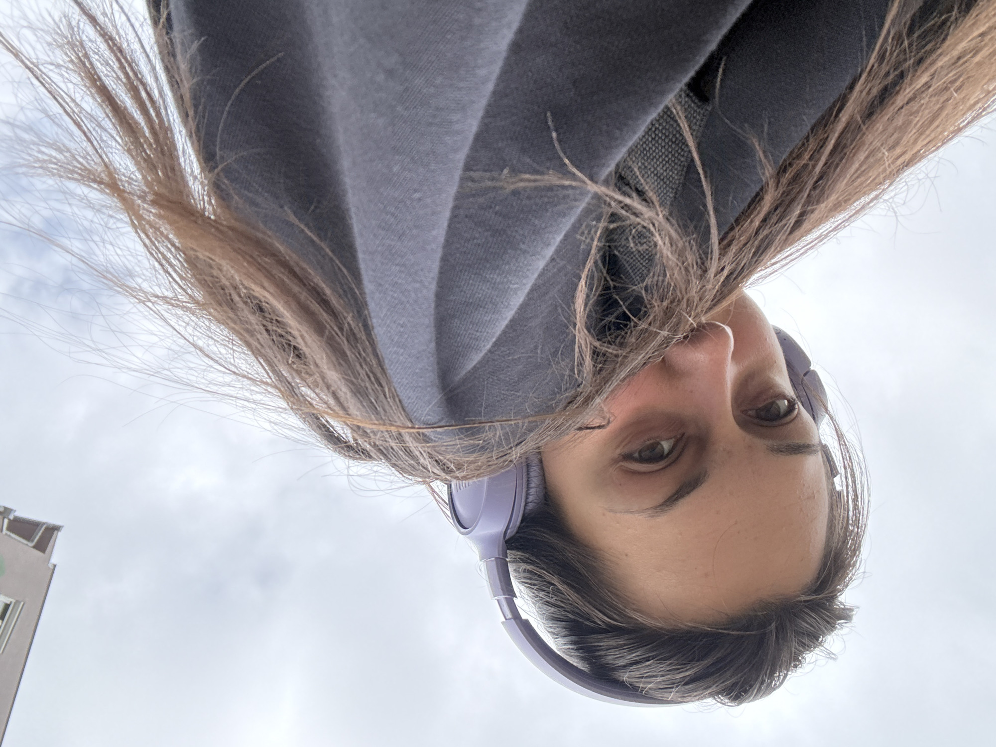 A low-angle shot shows a person with long, brown hair and light skin, wearing purple headphones, looking down at the camera. The person is wearing a dark grey hooded sweatshirt. Some of their hair is in front of the camera lens, partially obscuring the view. A building is faintly visible on the left side of the frame against an overcast sky. There are no other discernable elements to identify the location. No specific location in Kragujevac, Serbia can be determined.