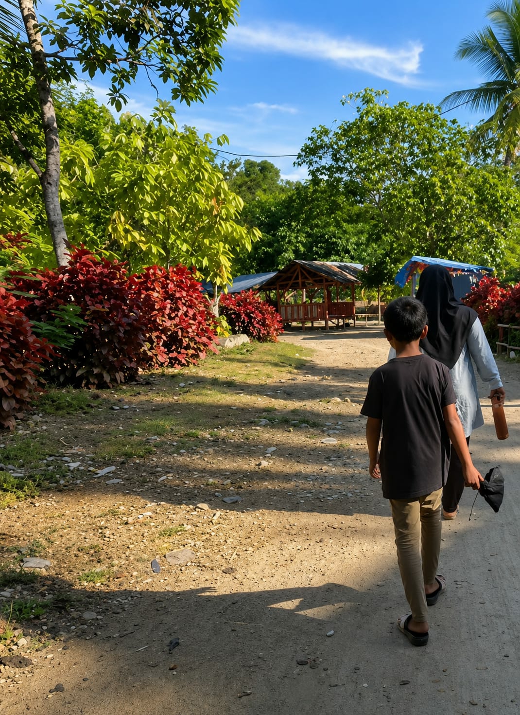 In Silaiyajae, Indonesia, a young boy wearing a black t-shirt and tan pants walks on a dirt path. Beside him, a woman in a light blue long-sleeved shirt and a black hijab walks, holding a brown bottle in her right hand and a black hat in her left. They are surrounded by lush greenery, including trees with vibrant green leaves and bushes with red foliage. In the background, there are two small wooden huts with red railings and blue roofs. The sky above is clear with a few white clouds, and the sunlight casts shadows on the ground, indicating it is daytime.