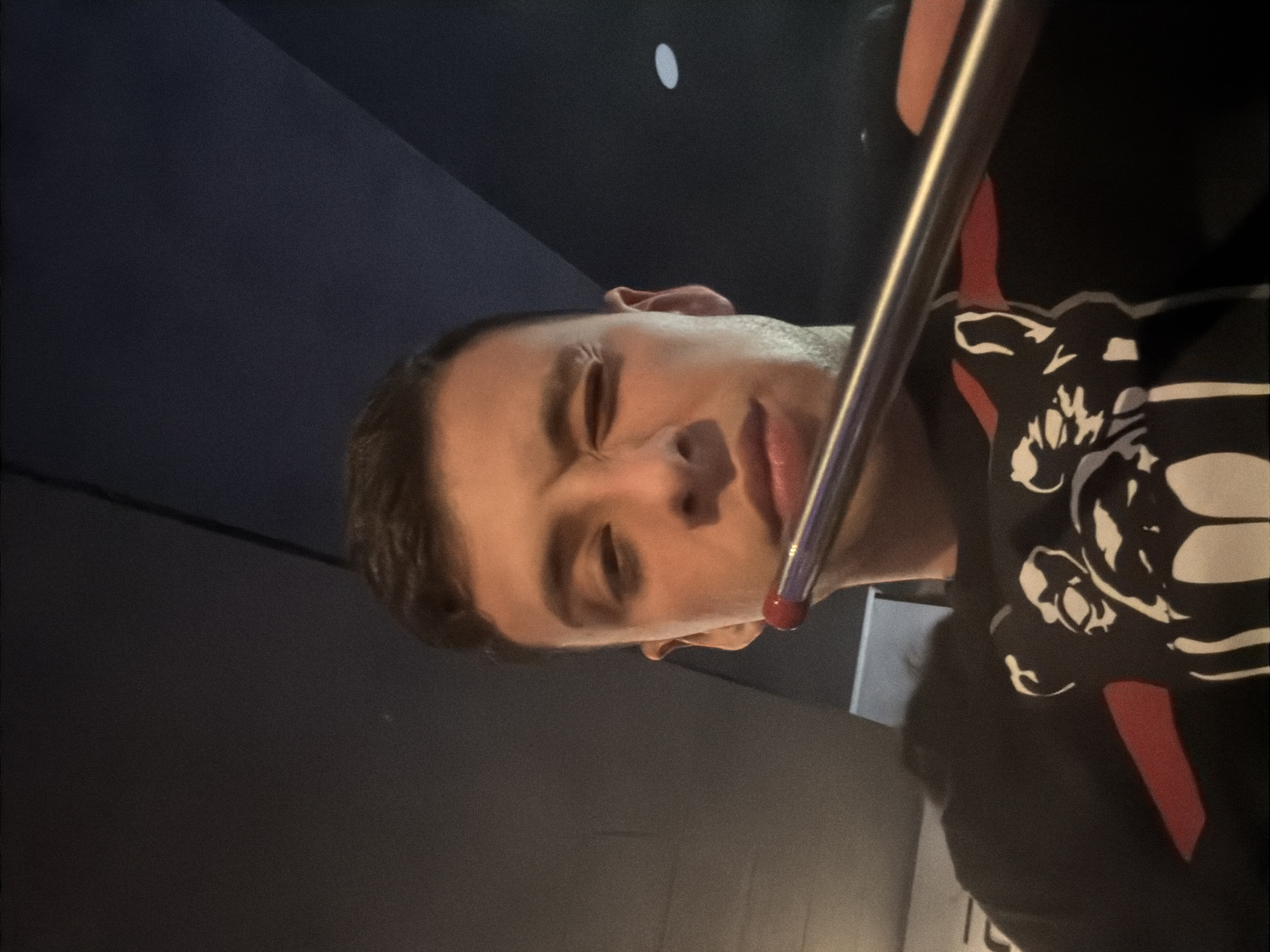 A close-up, dimly lit photograph features a young male subject with his head tilted horizontally across the frame. He has short dark hair and fair skin. His eyes are closed or heavily squinted, and his mouth is slightly open, giving him a relaxed or possibly drowsy expression.
A prominent, shiny metallic rod, possibly a straw or a vape pen, with a red tip is positioned near his mouth, extending diagonally across the image. He is wearing a dark-colored (likely black) hoodie or sweatshirt. The garment features a distinctive graphic design on the right side, consisting of abstract, stylized shapes in white and gray, resembling skulls or figures, complemented by a vertical red stripe or panel.
The background is dark and largely featureless, suggesting an indoor setting with low ambient lighting. A small, circular white light or reflection is faintly visible in the upper left background. The overall mood of the image is intimate and subdued, focusing on the subject and the object near his mouth. No visible text is present.