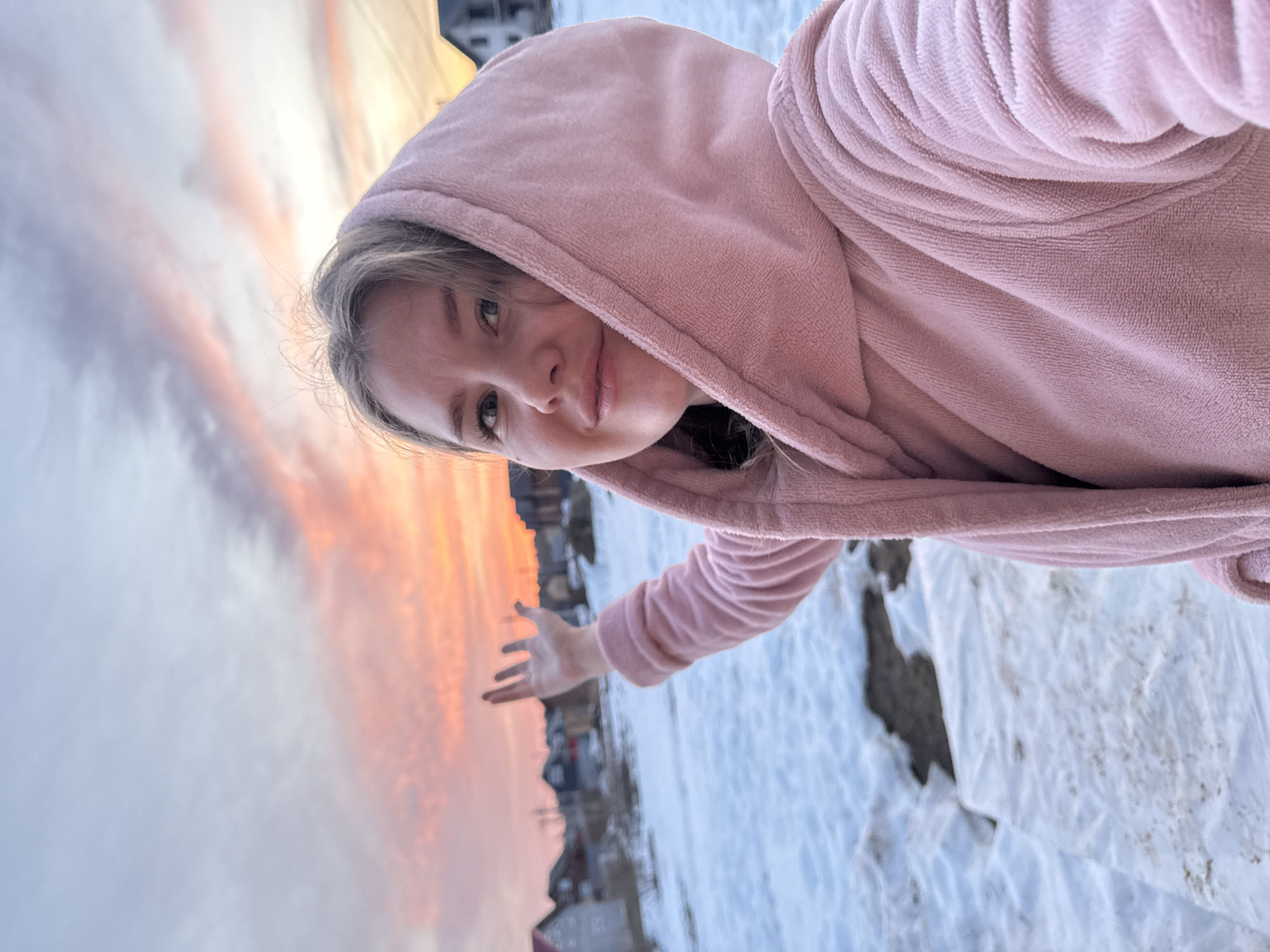 A woman with blonde hair and fair skin wearing a pink hooded robe is the primary subject of this rotated selfie-style photograph. She is situated outdoors in a snowy environment. Her right arm is extended towards the left of the frame, with her hand appearing to gesture towards the sky.
In the background, a vibrant sunset displays colors of orange and yellow. Below the horizon, there's a blurred view of what appears to be buildings. Patches of snow are visible on the ground. Based on location information provided, the setting is the city of Gornyi (Dvurechenskoe S/P), Russia.