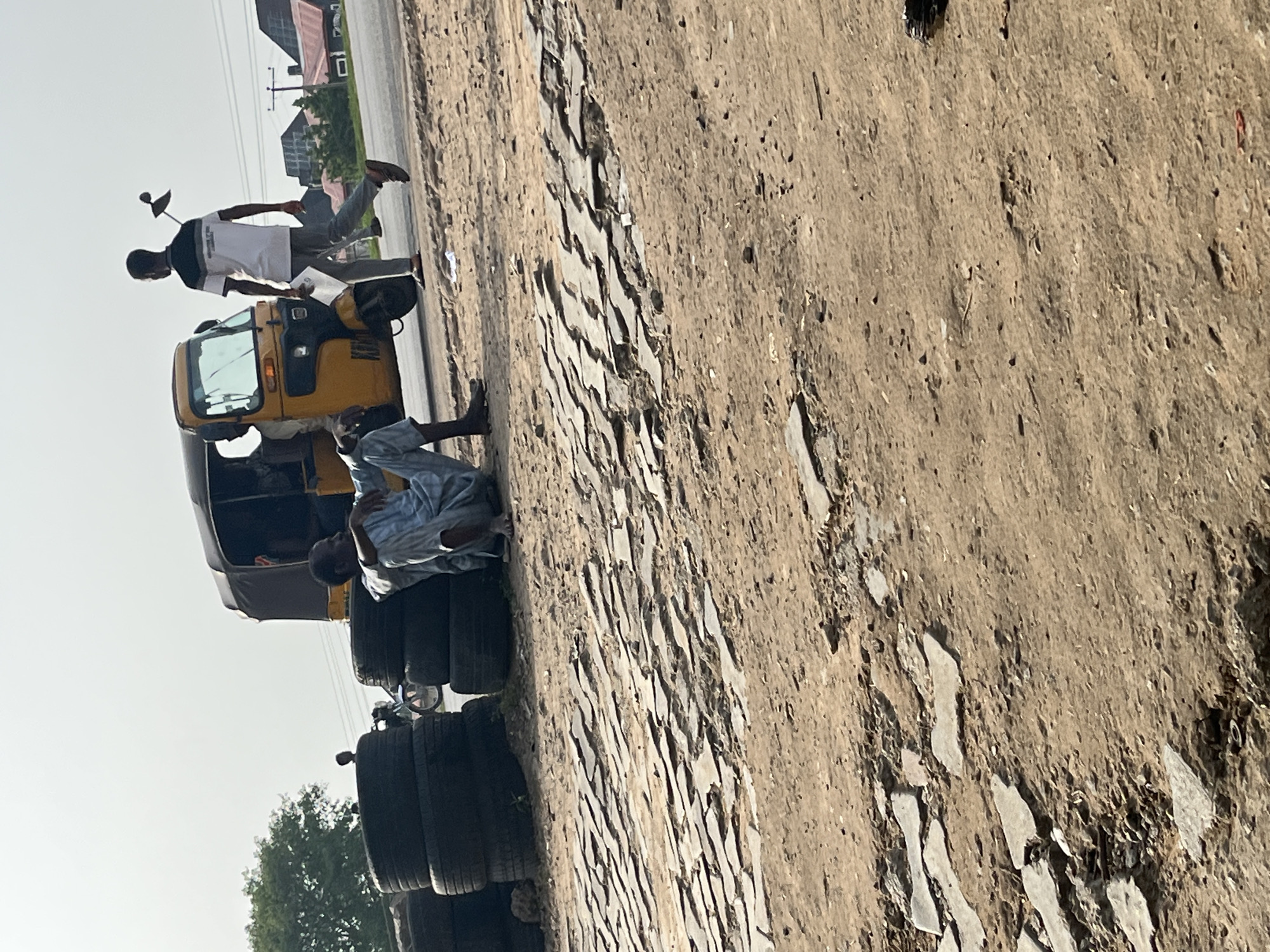 The image shows a scene in Abuja, Nigeria. A yellow auto rickshaw (tricycle) is parked on the side of a road next to a pile of tires. A man in a light blue robe is seated on the tires. Another man, dressed in a white shirt with dark sleeves and gray trousers, is walking away from the auto rickshaw while holding a piece of paper. The road surface is dirt and stone. In the background, there are buildings and power lines. The sky is bright.