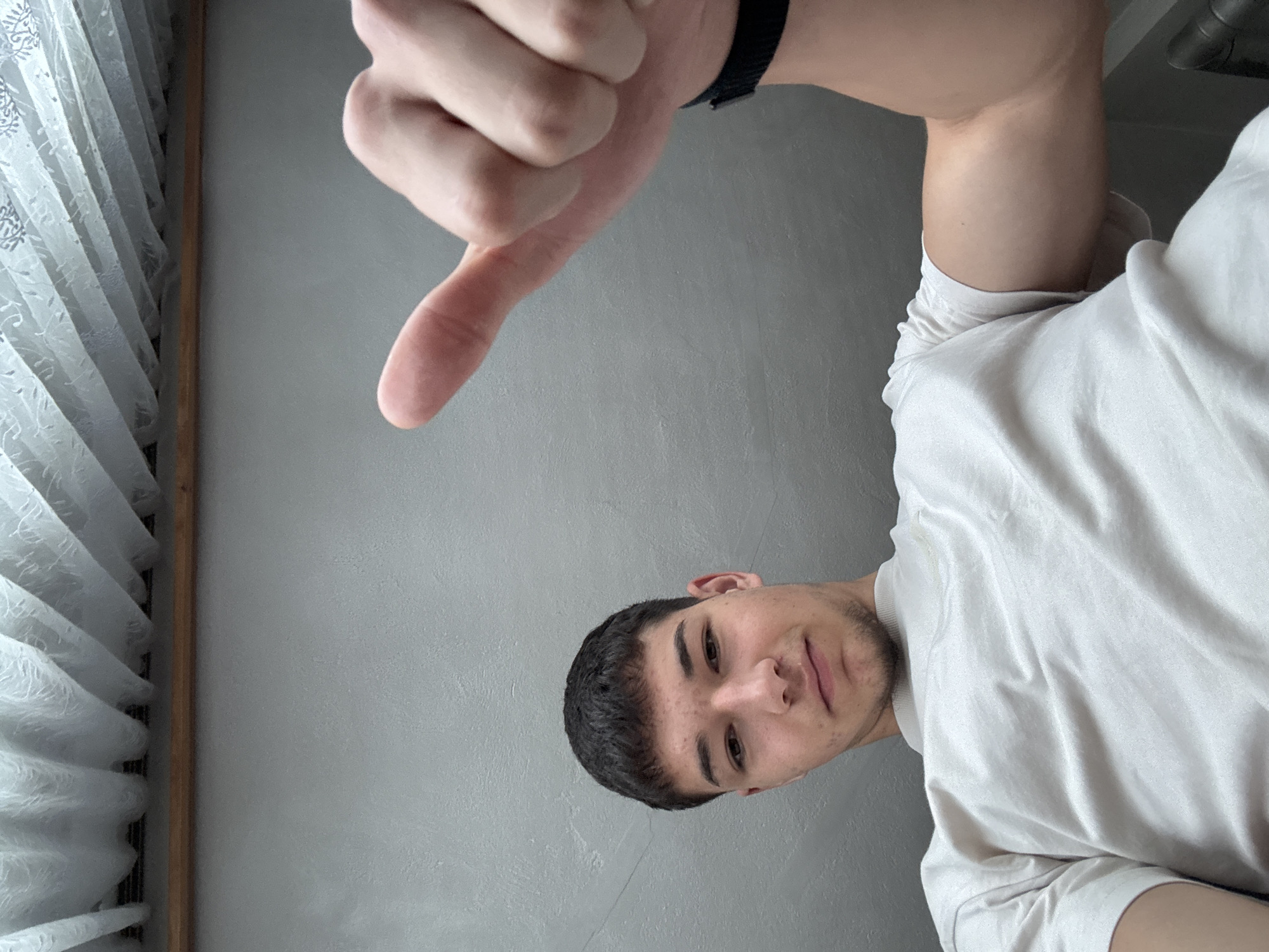 A young man is depicted in an indoor setting, possibly in Zlatograd, Bulgaria. The image is oriented with the man's head on the right side and his body extending to the left, suggesting a portrait taken at an unusual angle or rotated. He has short dark hair and a light beard, looking directly at the camera with a neutral to slightly serious expression. He is wearing a white t-shirt and a black band or watch on his right wrist. His right hand is prominently displayed in the upper center, making a "thumbs down" gesture. The background consists of a plain grey wall, and to the left, a window with white patterned curtains is visible, separated from the wall by a vertical wooden frame. The lighting appears to be bright and natural, indicating it is daytime. There is no visible text.
