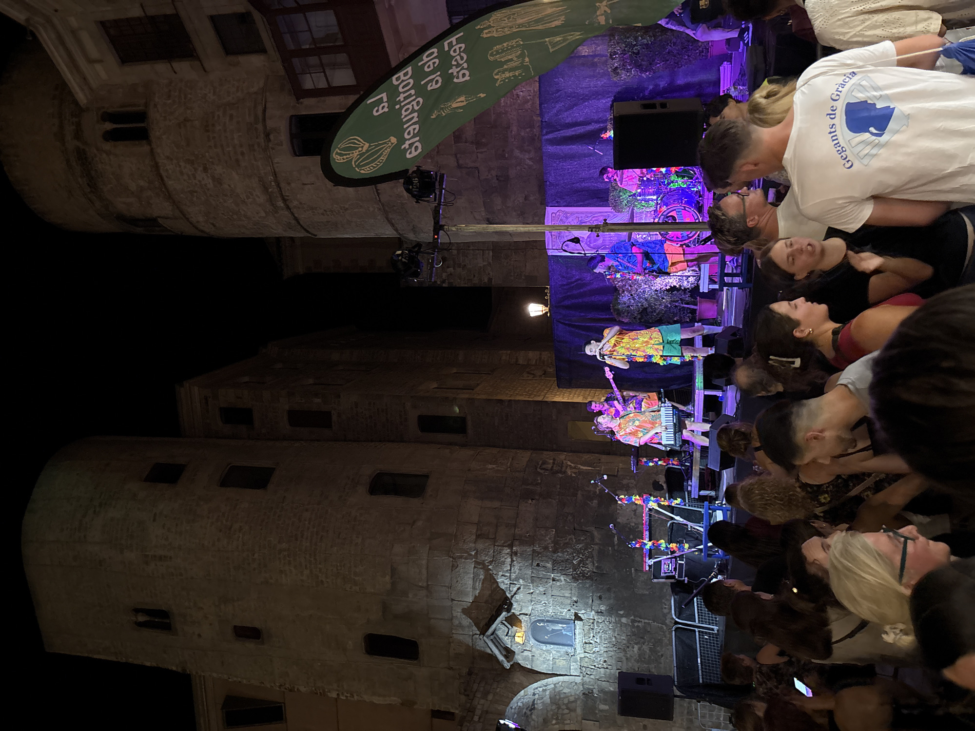 In Barcelona, Spain, a nighttime outdoor concert takes place in a historic plaza. The plaza is flanked by ancient stone buildings with arched windows. A stage is set up with musicians performing. A band, dressed in brightly colored Hawaiian shirts and shorts, plays instruments including keyboards, guitars, and drums. A large green banner with white text and illustrations hangs to the right of the stage. A crowd of people watches the performance, with some illuminated by stage lights and others in the shadows. The atmosphere appears festive and energetic.