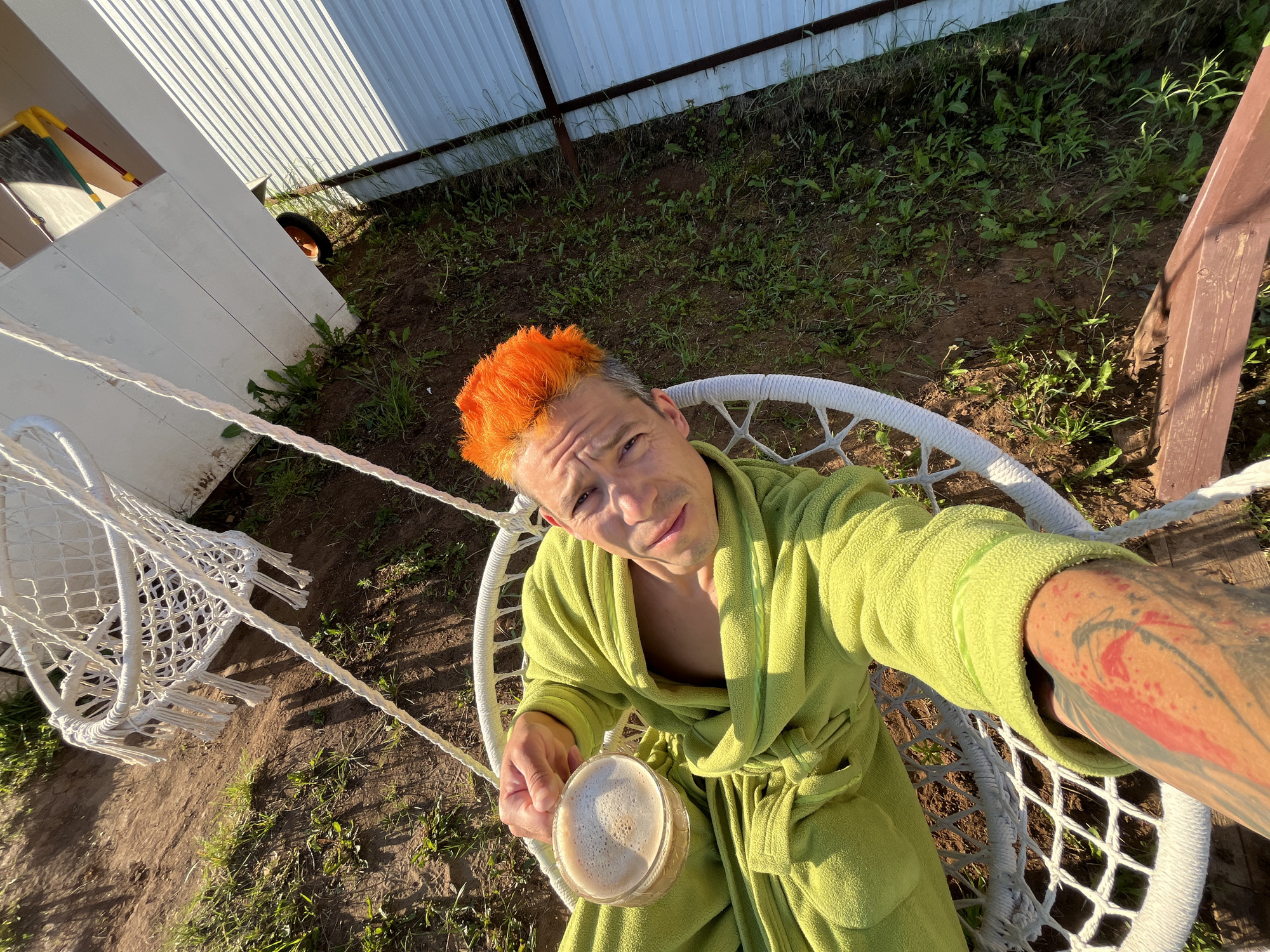 A man with brightly dyed orange hair, wearing a lime green bathrobe, sits in a white macrame hanging chair outdoors. He holds a clear glass mug of coffee in his left hand. His right arm, visible in the foreground, has a tattoo. The background shows a dirt and grassy area next to a white corrugated metal structure. The scene appears to be a backyard in Chaikovsky, Russia, with the sun casting shadows on the ground.