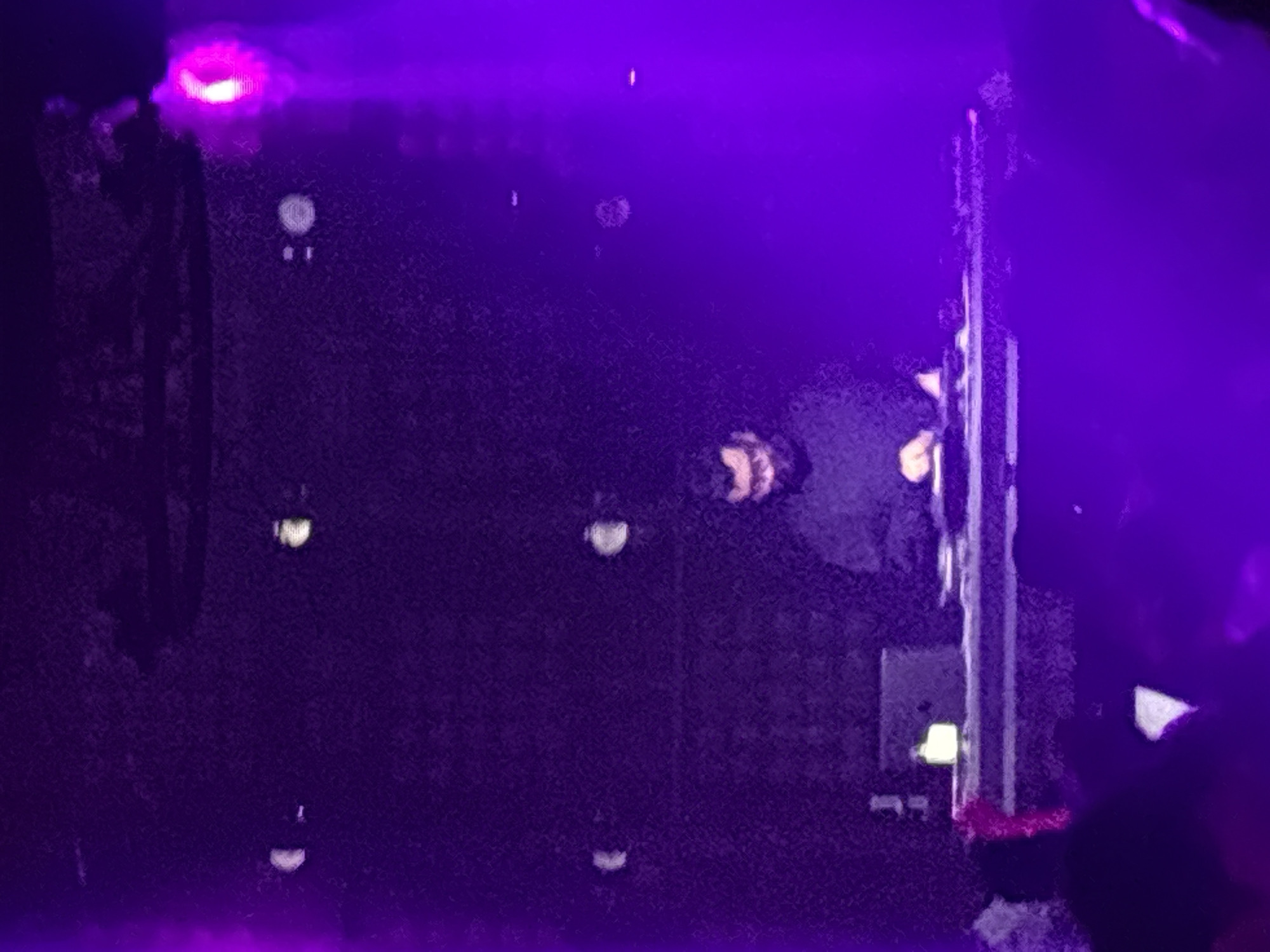 A dimly lit, purple-hued scene captures a person, likely a DJ, standing behind a console of electronic equipment on a stage. The DJ is wearing dark clothing and appears to be focused on the equipment. The console features illuminated buttons and what seems to be a laptop. The background is a textured dark surface, punctuated by several small, out-of-focus lights. The overall atmosphere suggests a live performance or event. The location is specified as North Jakarta, Indonesia.