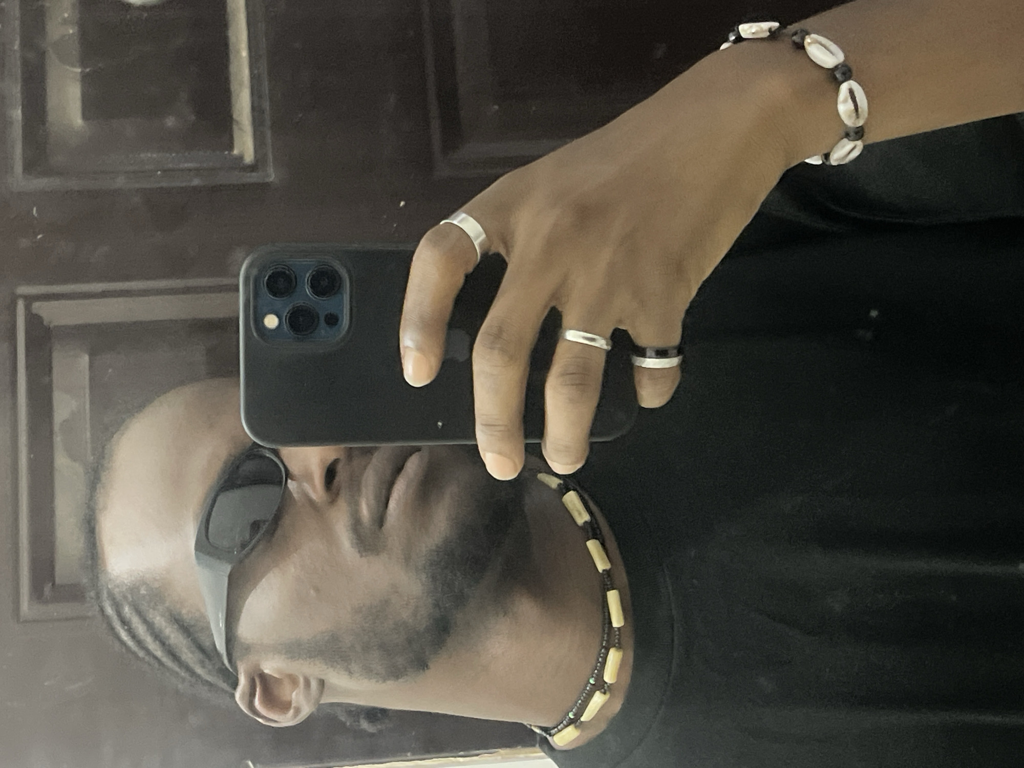 An individual with dark skin, located in the city of Jos, Nigeria, is captured in a mirror selfie. The person, whose gender appears male, wears dark, wraparound sunglasses that obscure the eyes. A cornrow hairstyle is visible on the sides of the head, complemented by facial hair consistent with a beard and mustache. The subject is dressed in a plain black t-shirt.
Around the neck, two layered necklaces are present: an inner necklace composed of small, dark beads, and an outer necklace featuring alternating light-colored, elongated rectangular beads and smaller dark beads. The individual's left hand, which holds a black smartphone (consistent with an iPhone model featuring a triple-lens camera array) for the photograph, displays three rings: a silver-toned band on the index finger, a silver-toned band on the middle finger, and a two-toned (black and silver-toned) wider band on the ring finger. The right wrist exhibits a bracelet composed of white cowrie shells interspersed with dark beads.
The background, observed through the mirror's reflection, consists of a dark, possibly wooden, wall or door featuring recessed rectangular panels. Minor smudges are visible on the mirror surface.
