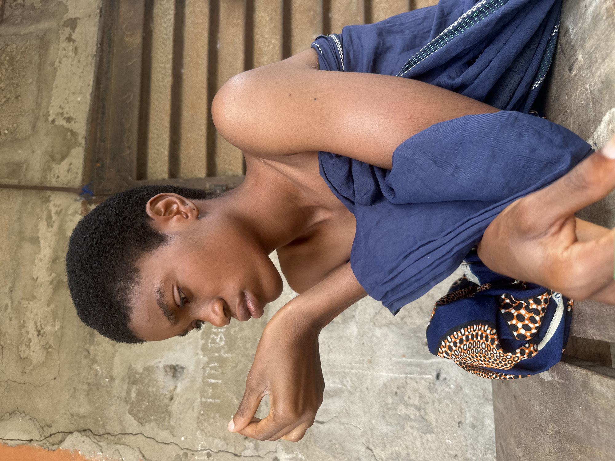 An individual with short, dark, curly hair is seated outdoors, facing to the left in profile. Their left arm is bent at the elbow, with the hand held near their face. They are draped in a dark blue fabric with a patterned trim featuring orange and white circular motifs. The background consists of a textured, weathered concrete wall with some faint markings, and wooden slatted doors or shutters. The person is positioned on a wooden surface, possibly a bench or table. The image appears to have been taken in Badagry, Nigeria, suggested by the specific style of the fabric and the architectural elements of the background, which are common in the region.