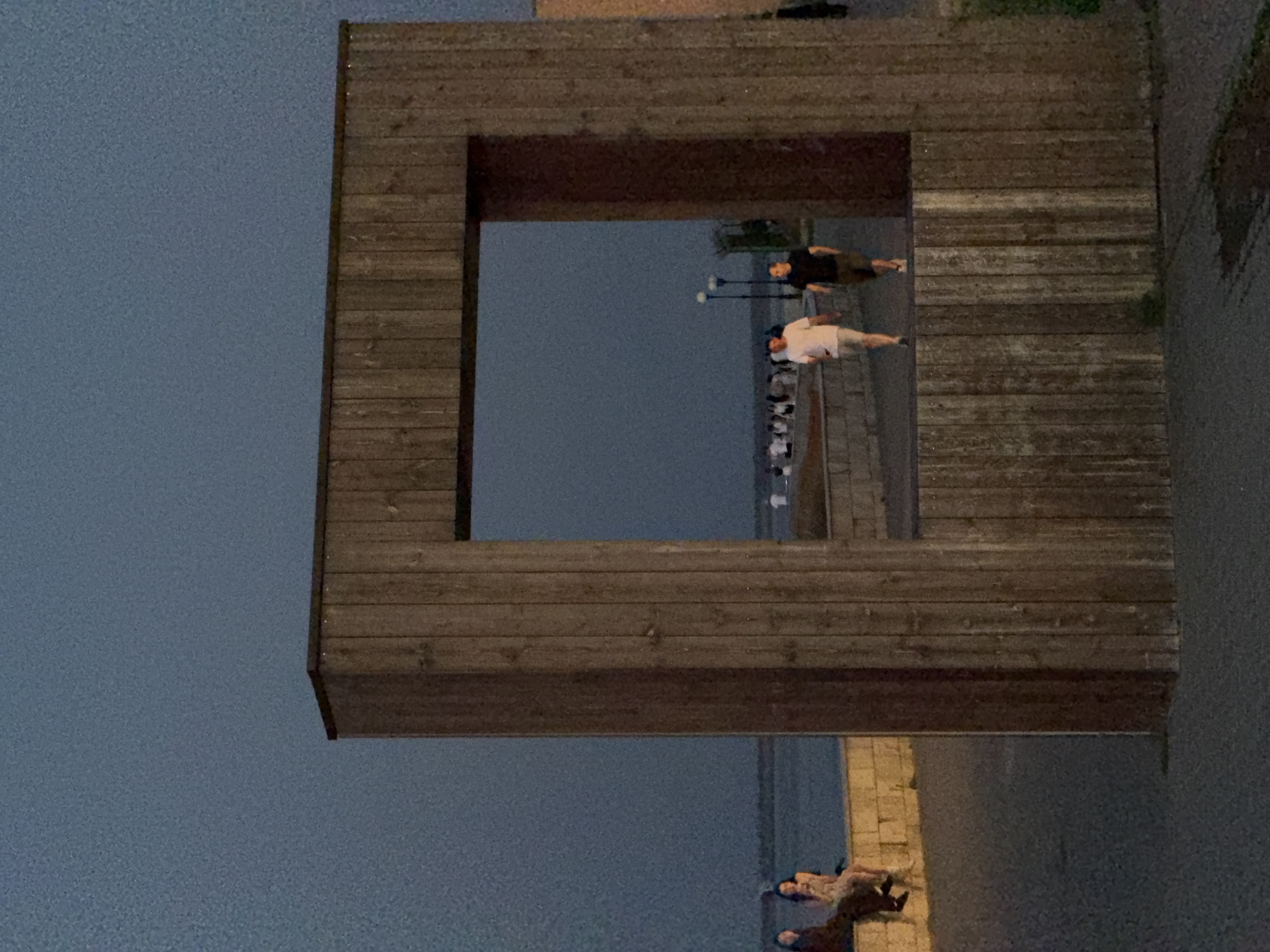 The image captures a twilight scene near the Volga River embankment in Kamyshin, Russia. A large, square wooden frame structure dominates the foreground, its vertical planks weathered and aged. Through the frame, a paved walkway is visible, bordered by a low stone wall.
On the left side of the walkway, two young women are seated on the stone wall, facing the water. One is wearing a light-colored dress, and the other is in darker clothing. Further down the walkway, within the frame, two men are walking. The man on the left wears a white t-shirt and shorts, while the man on the right is dressed in a black t-shirt and shorts. In the background, beyond the men, a group of people are visible strolling along the embankment, with streetlights illuminating the path. The sky is a deep blue, characteristic of dusk, and the water of the Volga River is calm. The overall impression is one of a peaceful evening by the river.