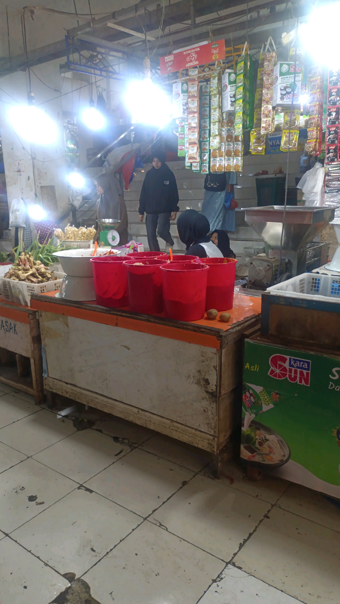 The image depicts an indoor market scene, likely in Padang Panjang, Indonesia, characterized by bright overhead lighting and the presence of various goods and people. In the foreground, a wooden counter displays several large red buckets, some filled with what appears to be food items, and a white bowl. To the left of the counter, a crate of ginger and other produce is visible. Behind the counter, several individuals are engaged in market activities. One person in a black hijab and jacket stands near the counter, while another in a grey hijab and dress is seen by a green scale. Further back, a person in a blue garment is partially visible, and a person in a black hijab is seated. The background shows stacked shelves with packaged goods, suggesting a retail environment. The floor is tiled and appears somewhat worn. A prominent green advertisement for "Kara SUN" with an image of a food bowl is affixed to the side of a structure in the lower right portion of the frame.