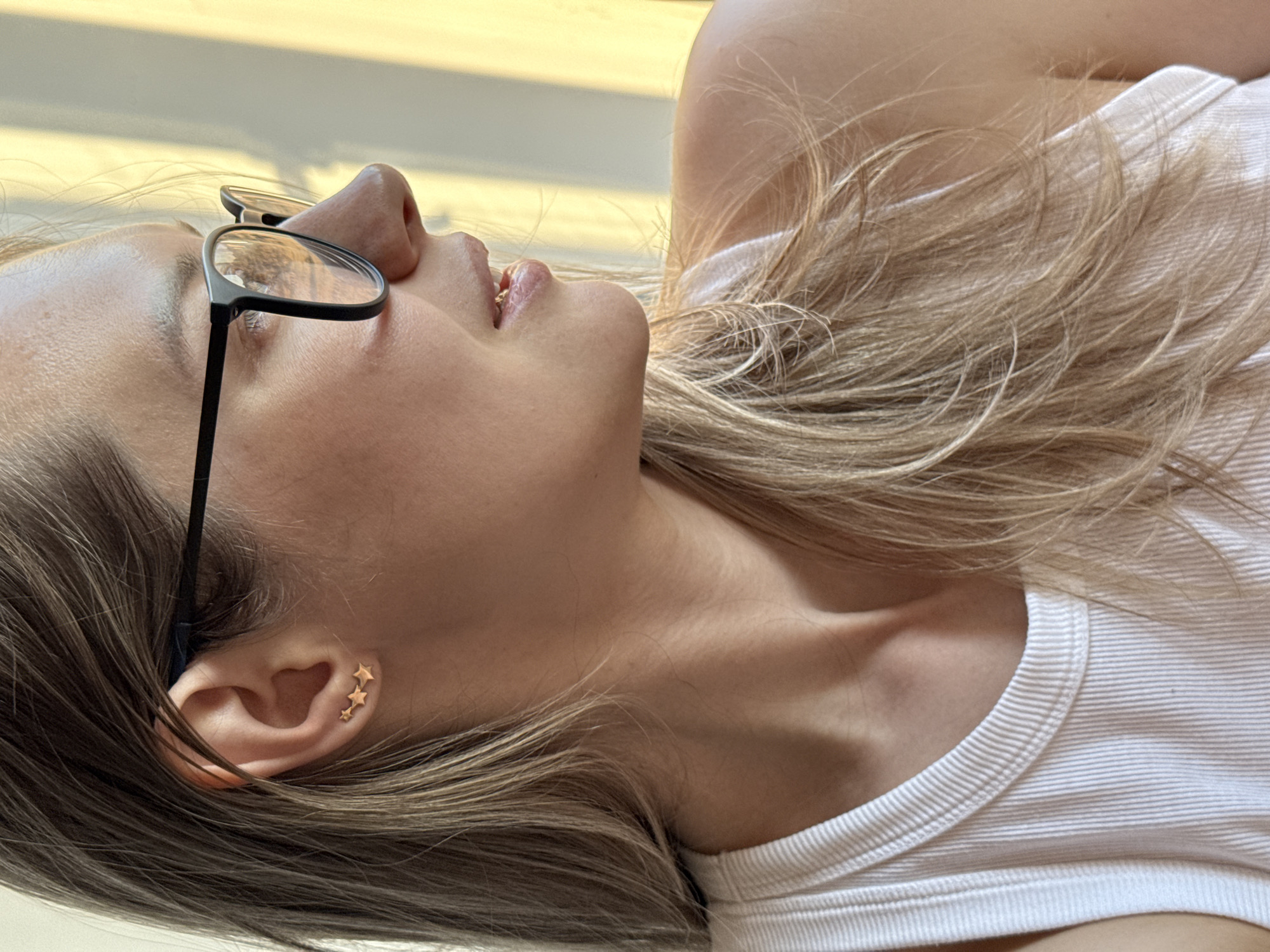 A close-up, side-profile shot of a fair-skinned woman wearing black-framed glasses, a white ribbed tank top, and three star-shaped earrings in her left ear. The woman has light brown hair. The background appears to be a blurry, light-colored surface, possibly a wall or ceiling. There is no indication of Chaikovsky, Russia in the image.