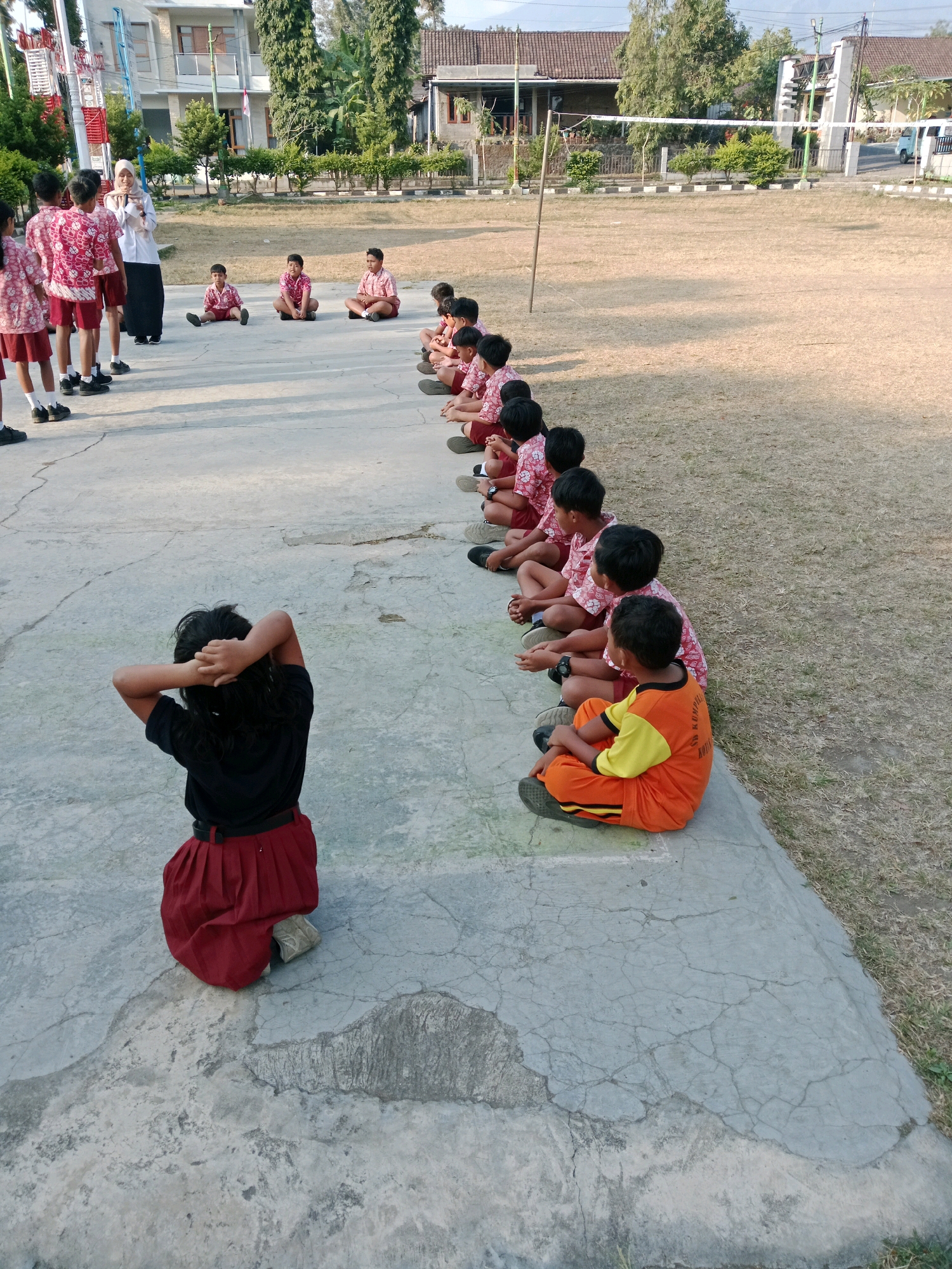 In Salatiga, Indonesia, a group of schoolchildren in uniform are sitting in rows on a concrete surface next to a grassy field. The students, primarily boys, wear red and white patterned shirts and dark shorts or pants, while a few girls wear red skirts. In the foreground, a student with dark hair is facing away from the camera, with their hands behind their head. Another student in an orange and yellow outfit sits to the right. A woman in a hijab and white top is standing and addressing the children. Behind them, there are buildings and trees. The scene appears to be outdoors, possibly at a school during an activity or lesson.