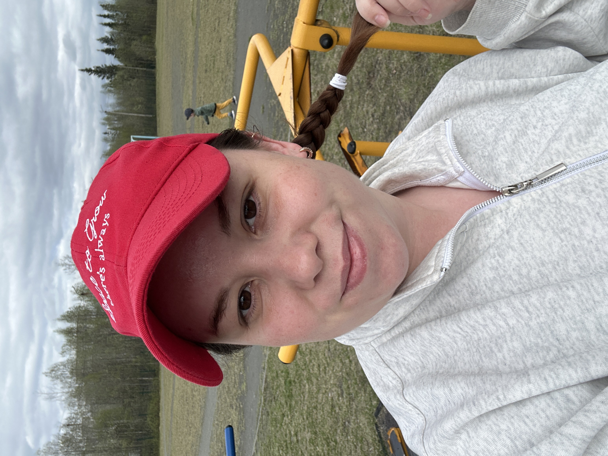 A woman wearing a red baseball cap and a gray zip-up sweatshirt is standing outside. The woman has brown hair in a braid with white elastics. The setting appears to be a park with yellow playground equipment, grass, trees, and a cloudy sky. A person is in the background, in the distance.